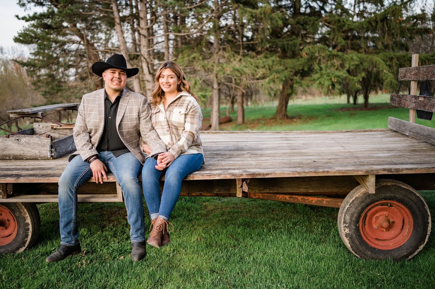 A couple in casual western wear sits together on an old wooden farm wagon.