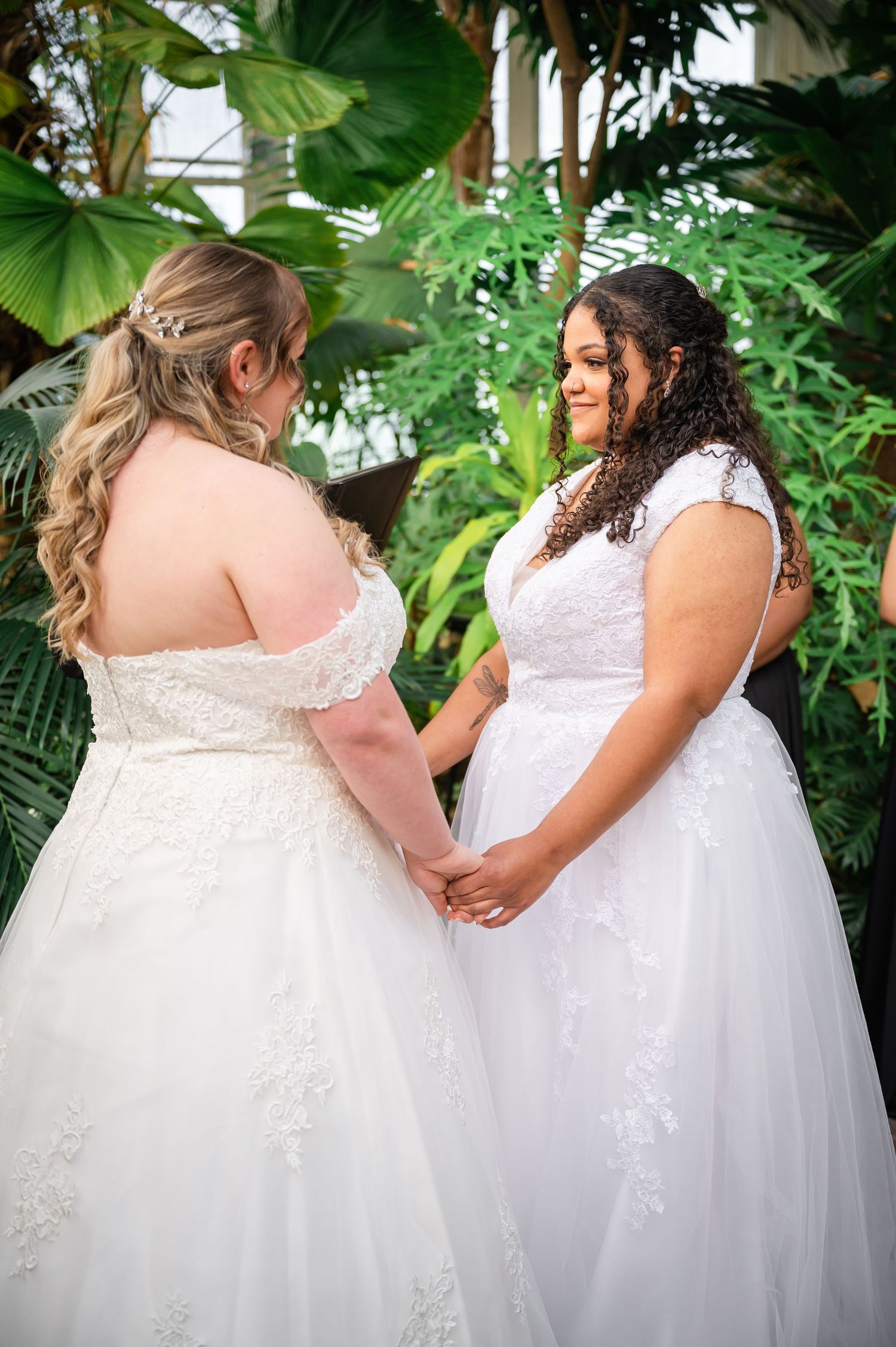 Two people in white wedding dresses hold hands in front of tropical greenery background.