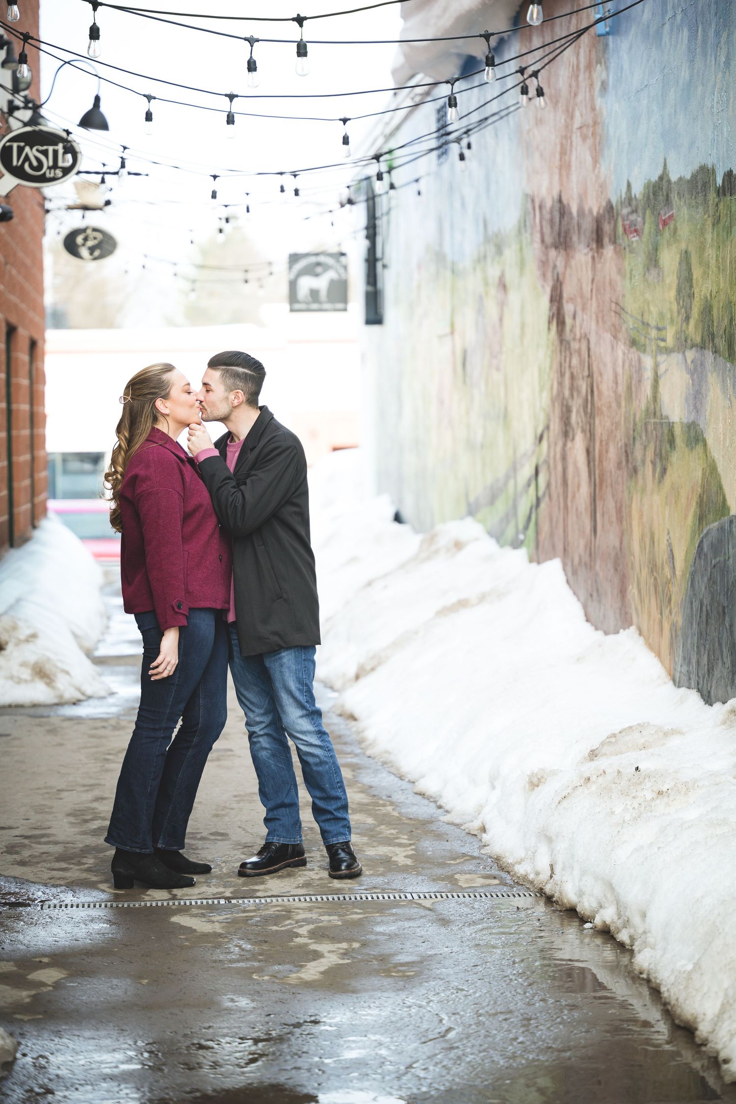 A romantic winter engagement photo session showing a couple sharing tender moments along a snowy alleyway with string lights above.