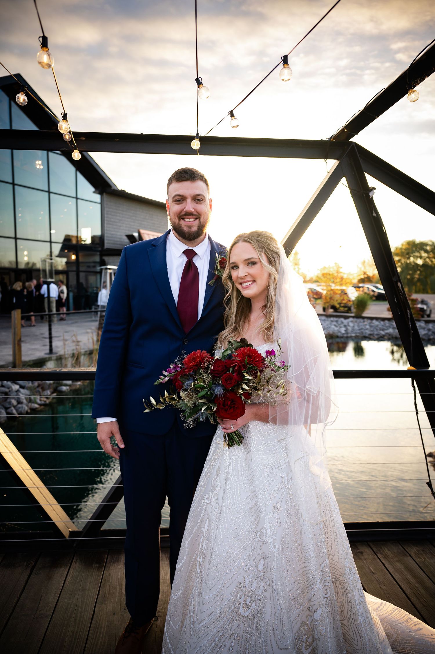 Wedding portrait on deck with string lights and glass building backdrop at golden hour.