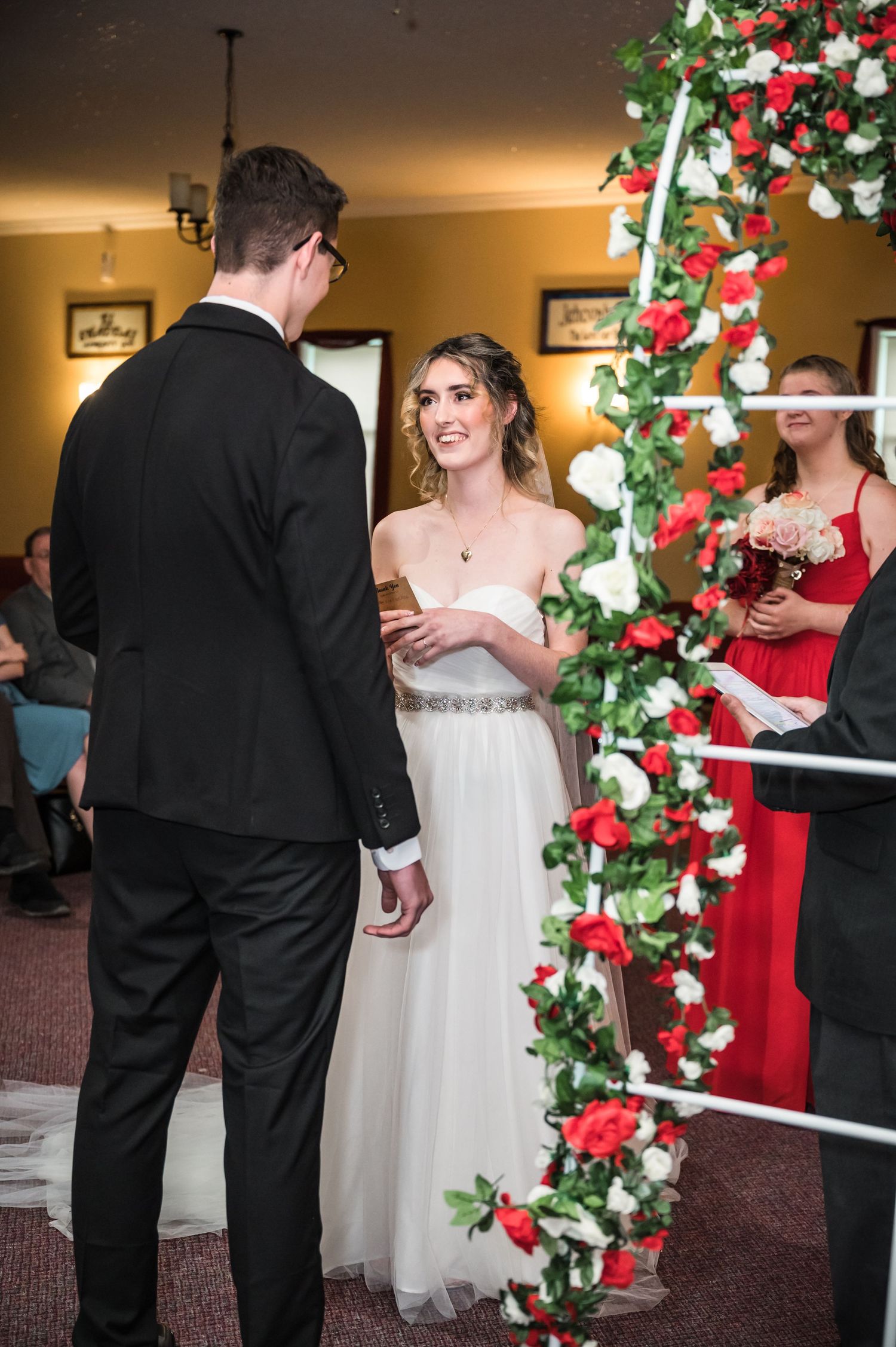 Indoor wedding ceremony moment near decorated floral arch with red and white flowers.