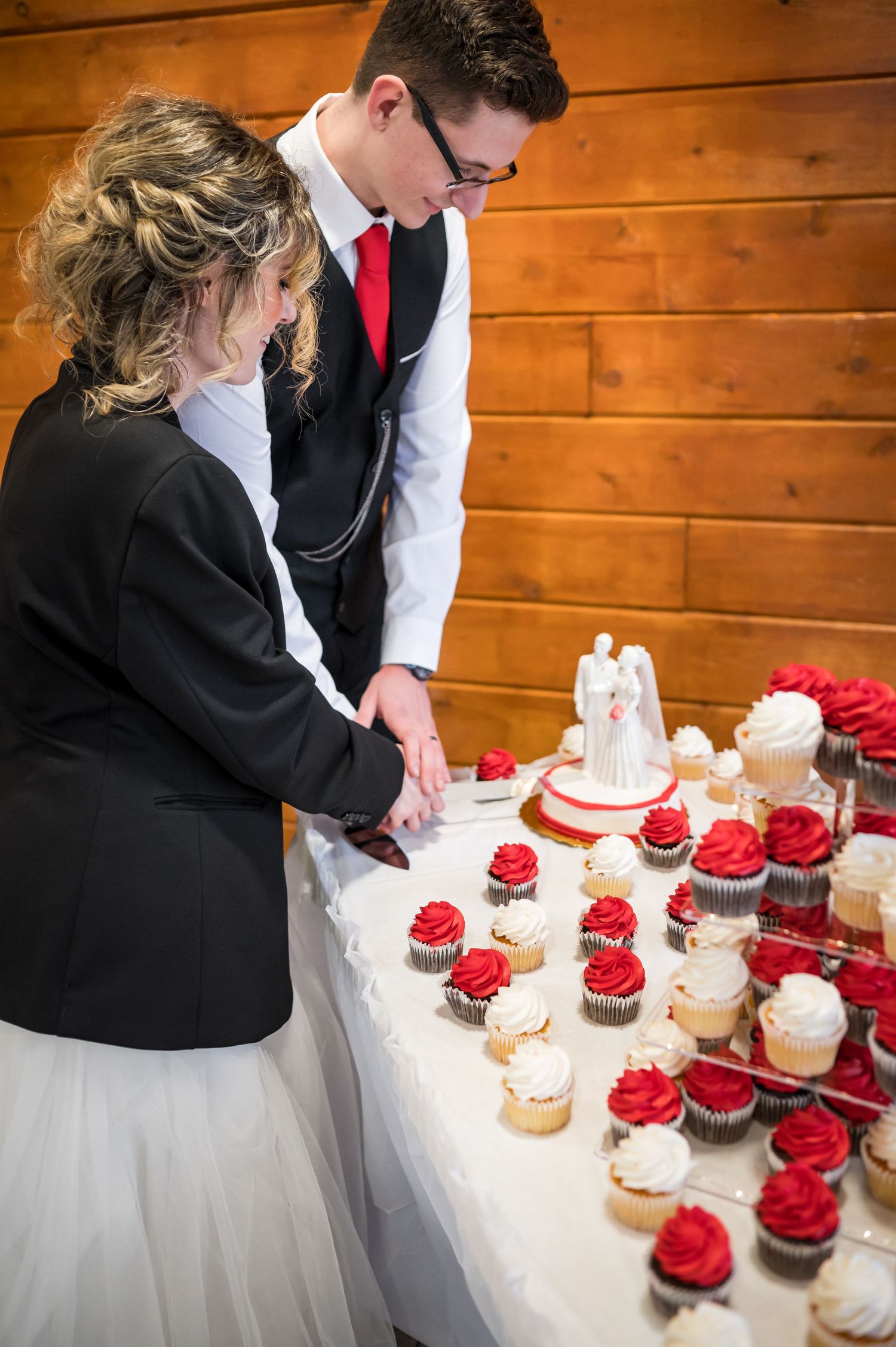 Wedding couple cuts cupcakes at rustic wooden reception venue with red and white dessert display.