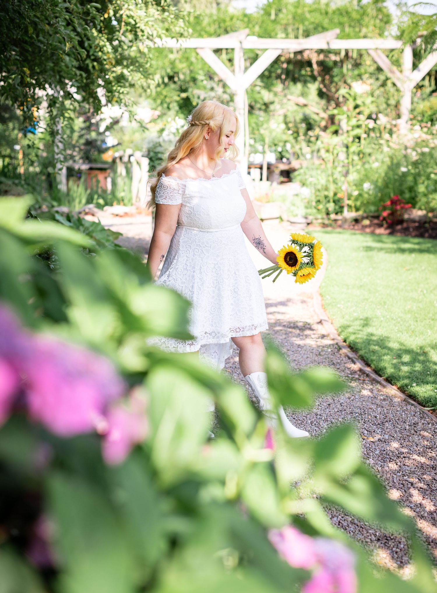 Sunflower bouquet and white lace dress create a romantic garden scene framed by pink flowers.