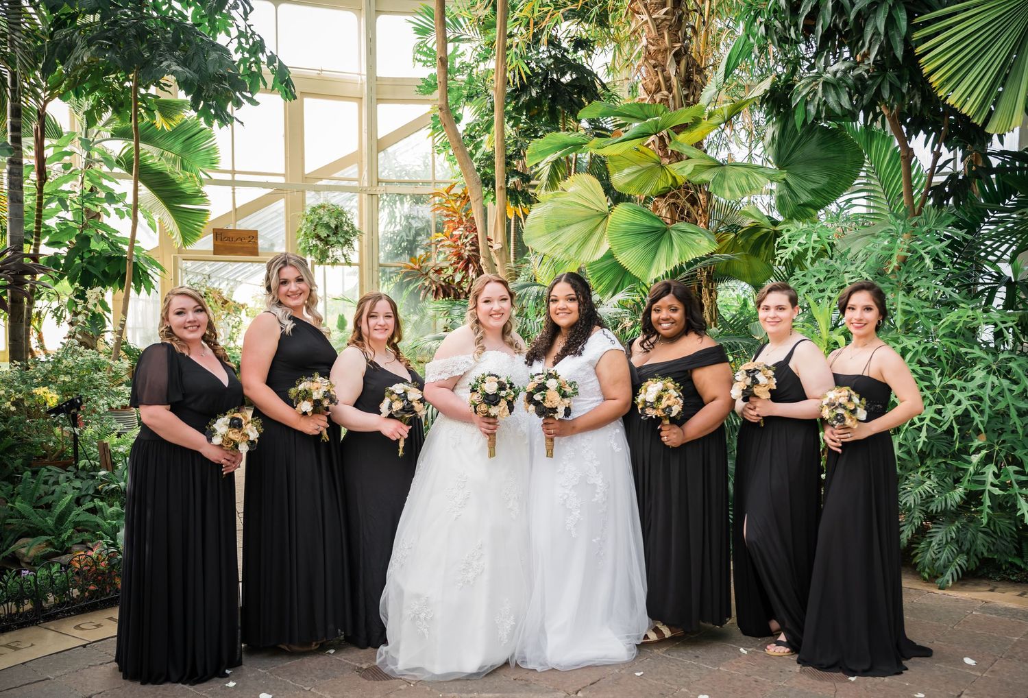 Wedding party wearing black dresses and white gowns pose together in a tropical greenhouse setting.