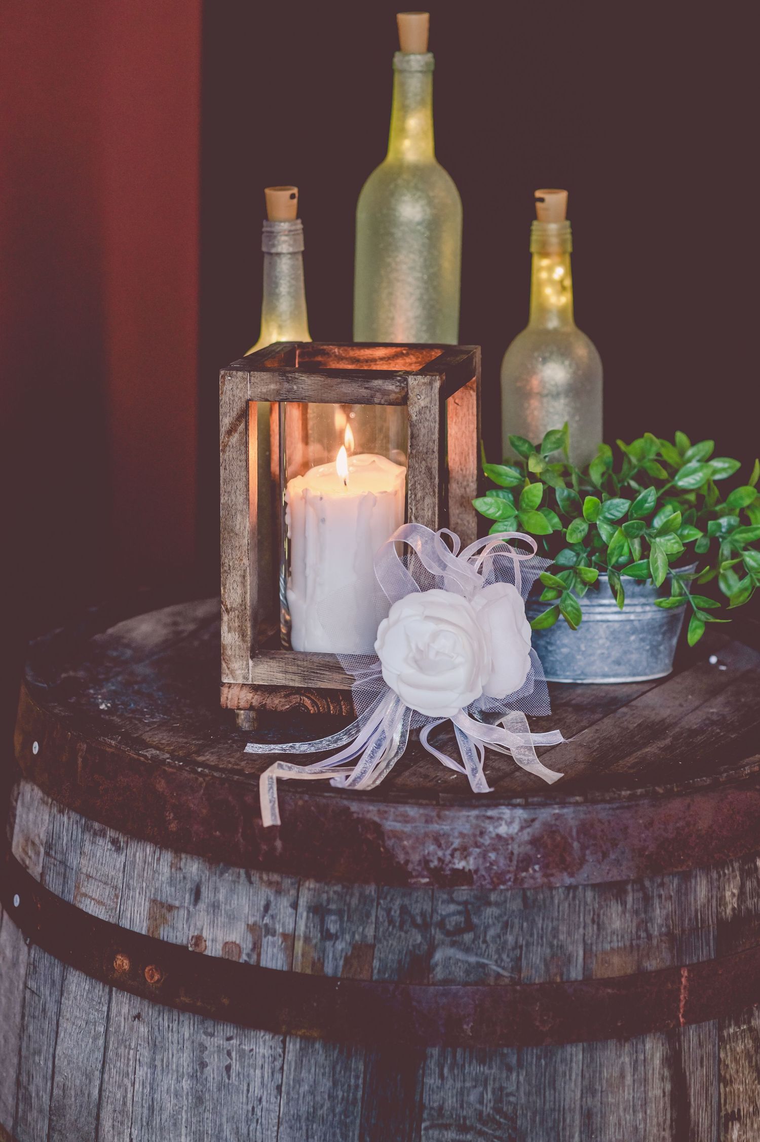 Wedding decor featuring illuminated bottles, a candle in a rustic lantern, and greenery on a wooden barrel.