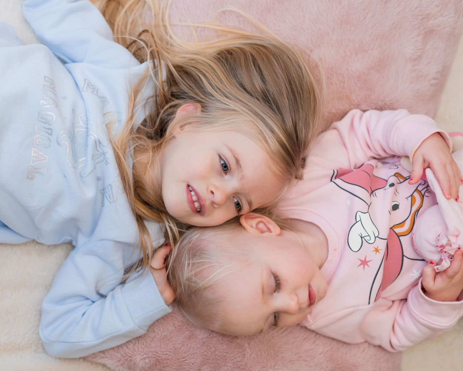 Two children resting together on pink bedding while wearing matching blue and pink tops.