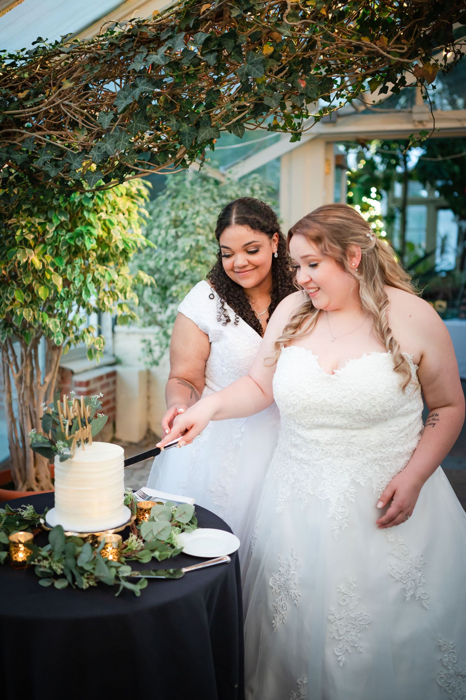 Two brides in elegant white gowns share a moment while cutting their wedding cake under garden greenery.
