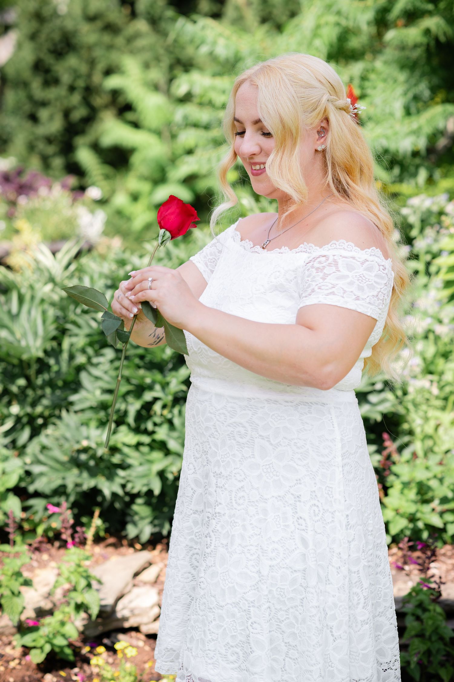 Garden portrait with white lace dress and red rose against green foliage.