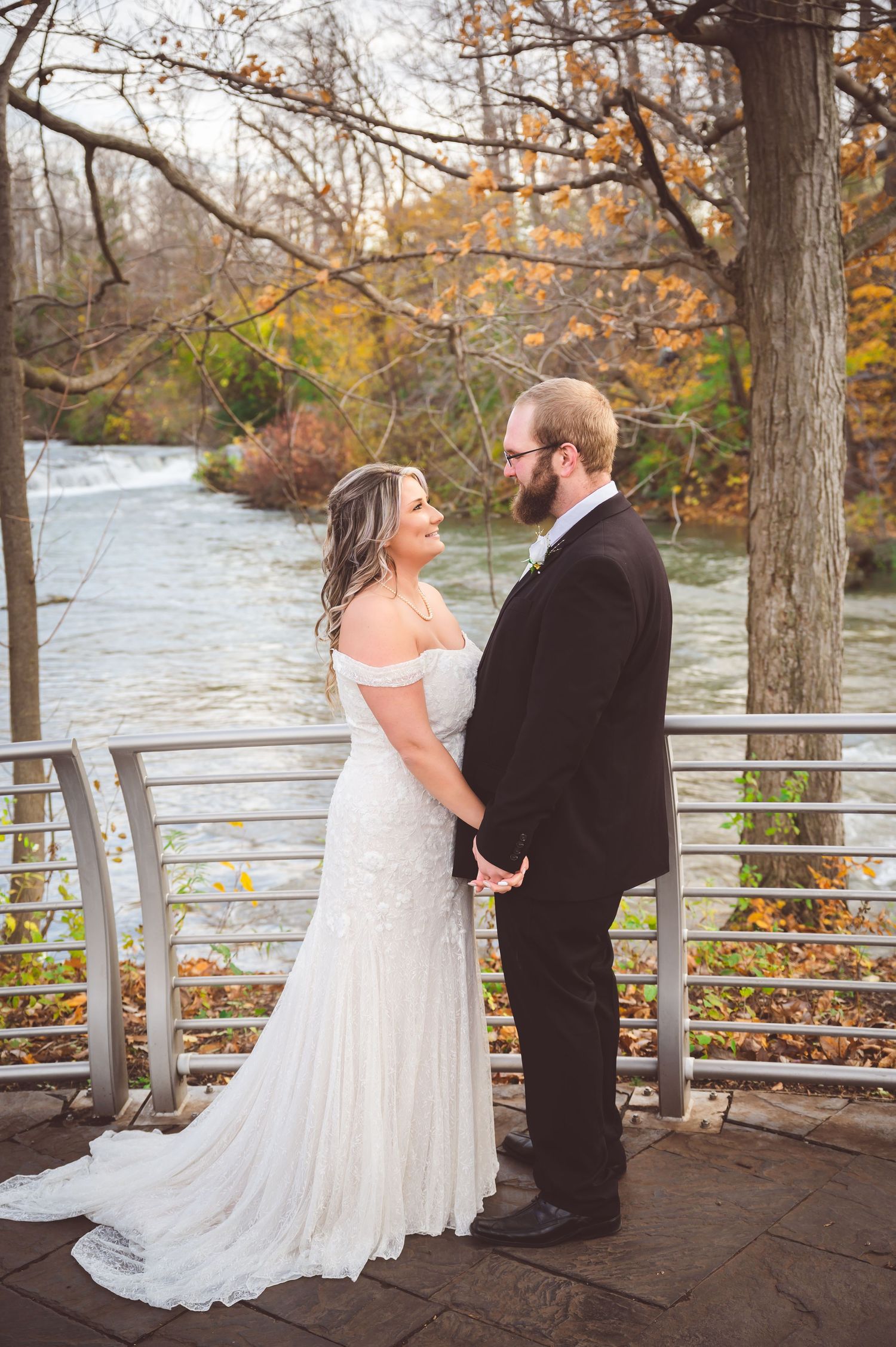 A wedding couple shares a romantic moment on a riverside deck during autumn with fall foliage in the background.