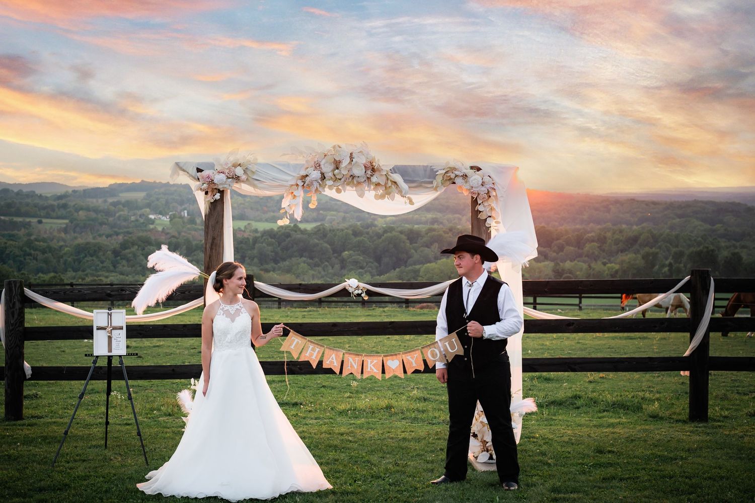 Scenic sunset wedding photo with flowing veil against mountain landscape and wooden fence.