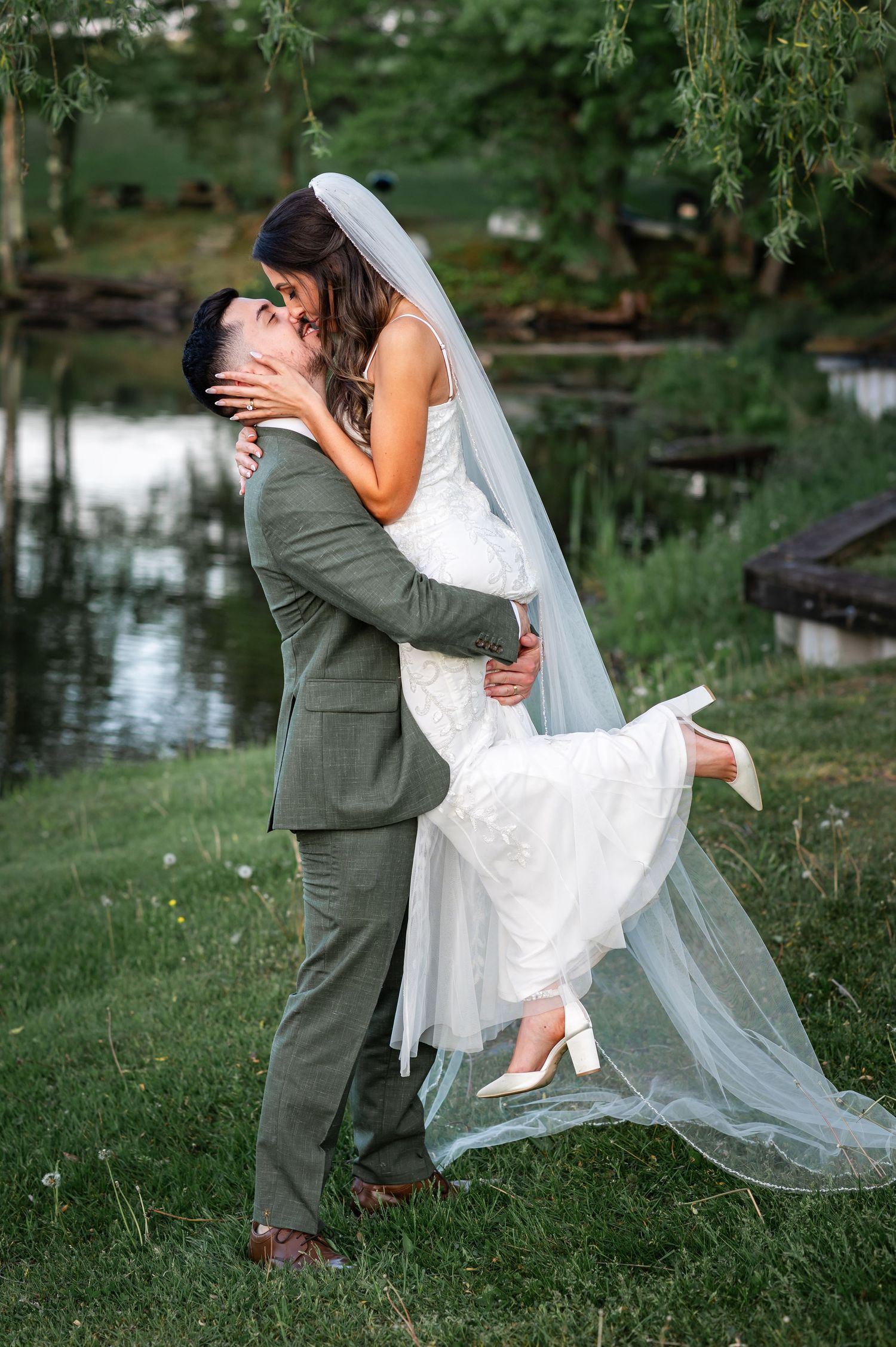 Newly married couple shares romantic kiss by a reflective pond with willow trees and flowing white veil in the background.