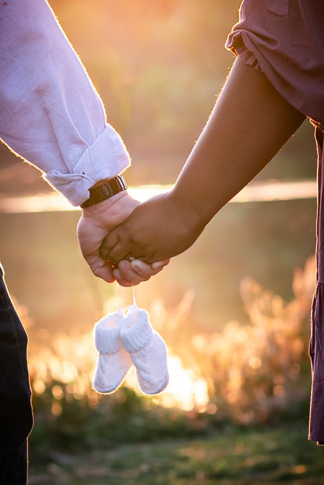 Couple holding hands with tiny baby shoes dangling between them at sunset.