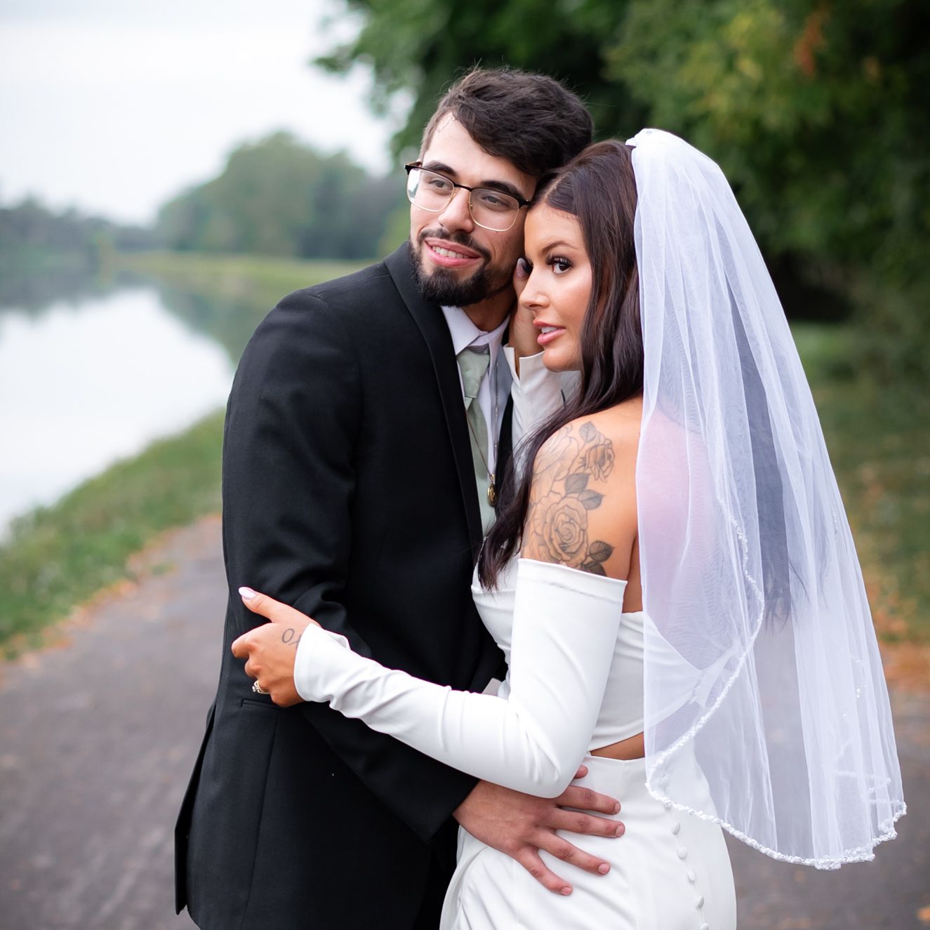 Newlywed couple embraces on a path by a peaceful lake during their countryside wedding photoshoot.