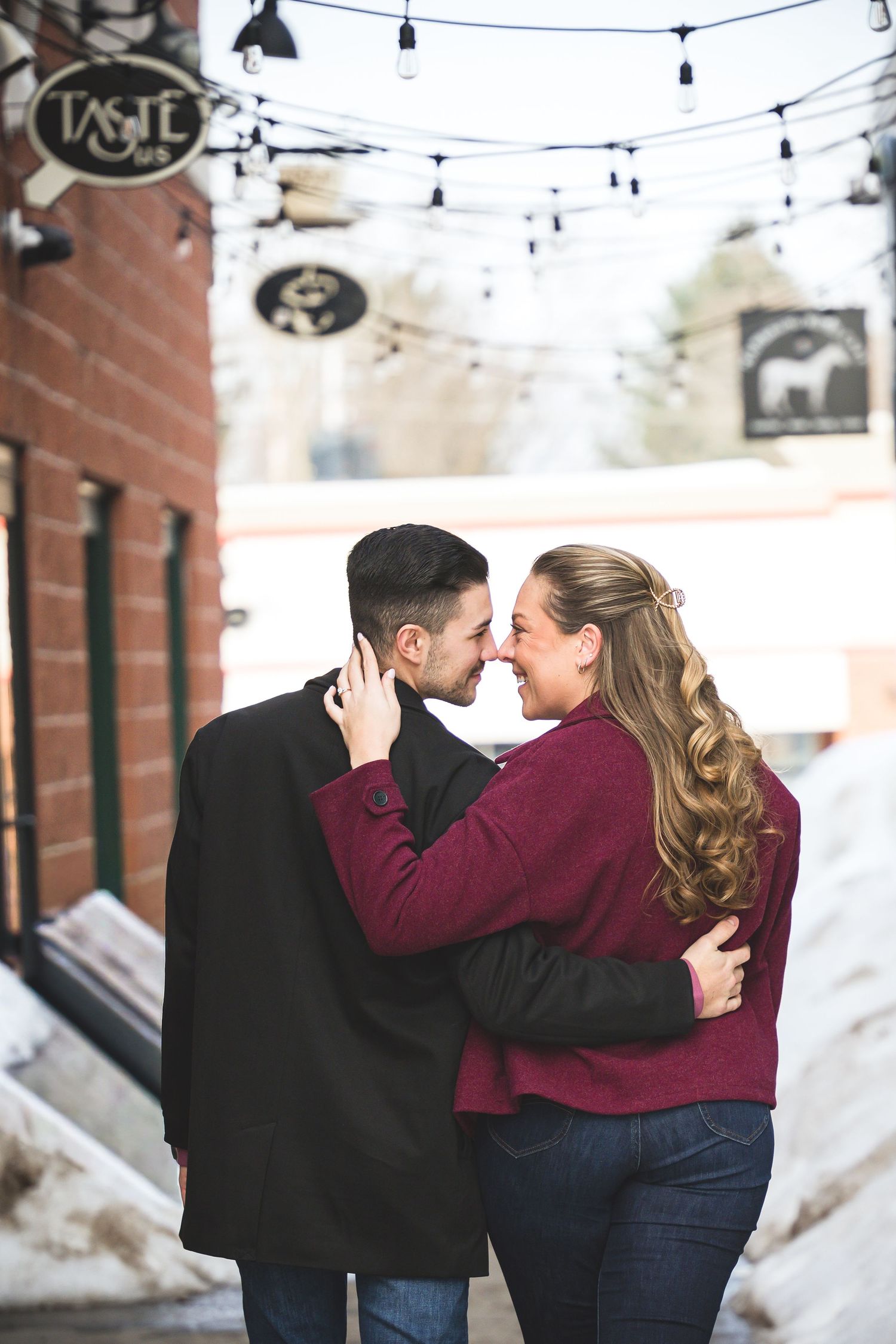 Couple embracing and laughing together on a snowy street with string lights overhead near a brick building.