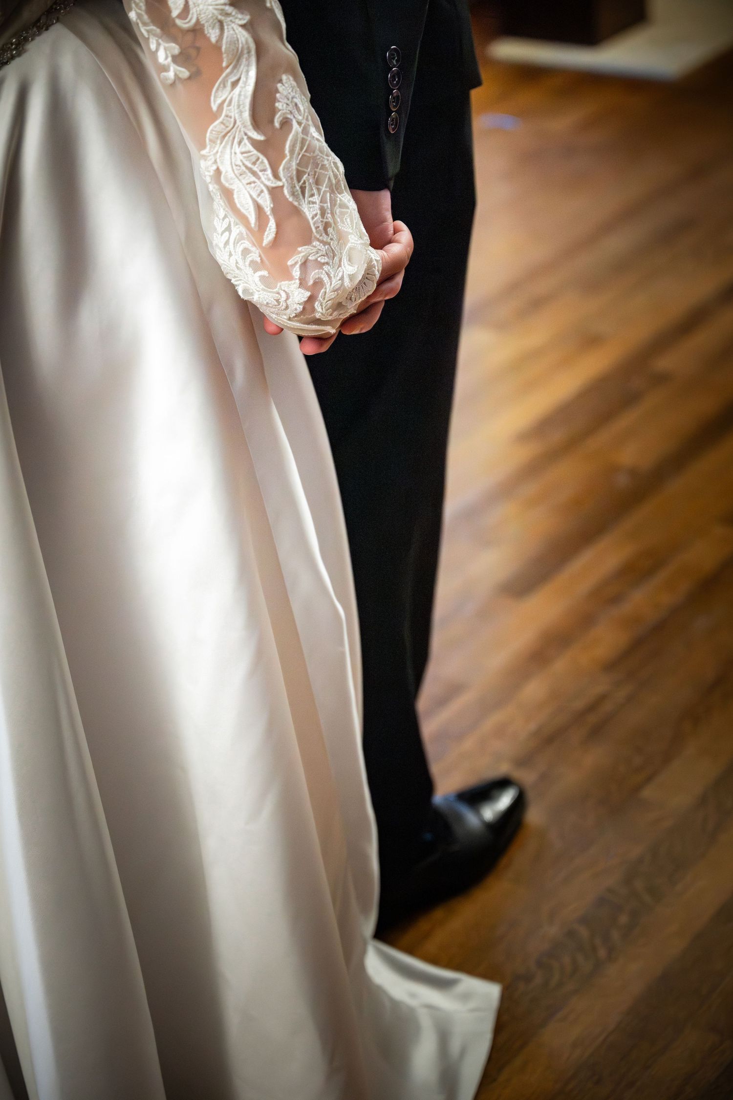 Close-up of a bride and groom standing together on hardwood floor showing detailed lace sleeve and formal attire details.