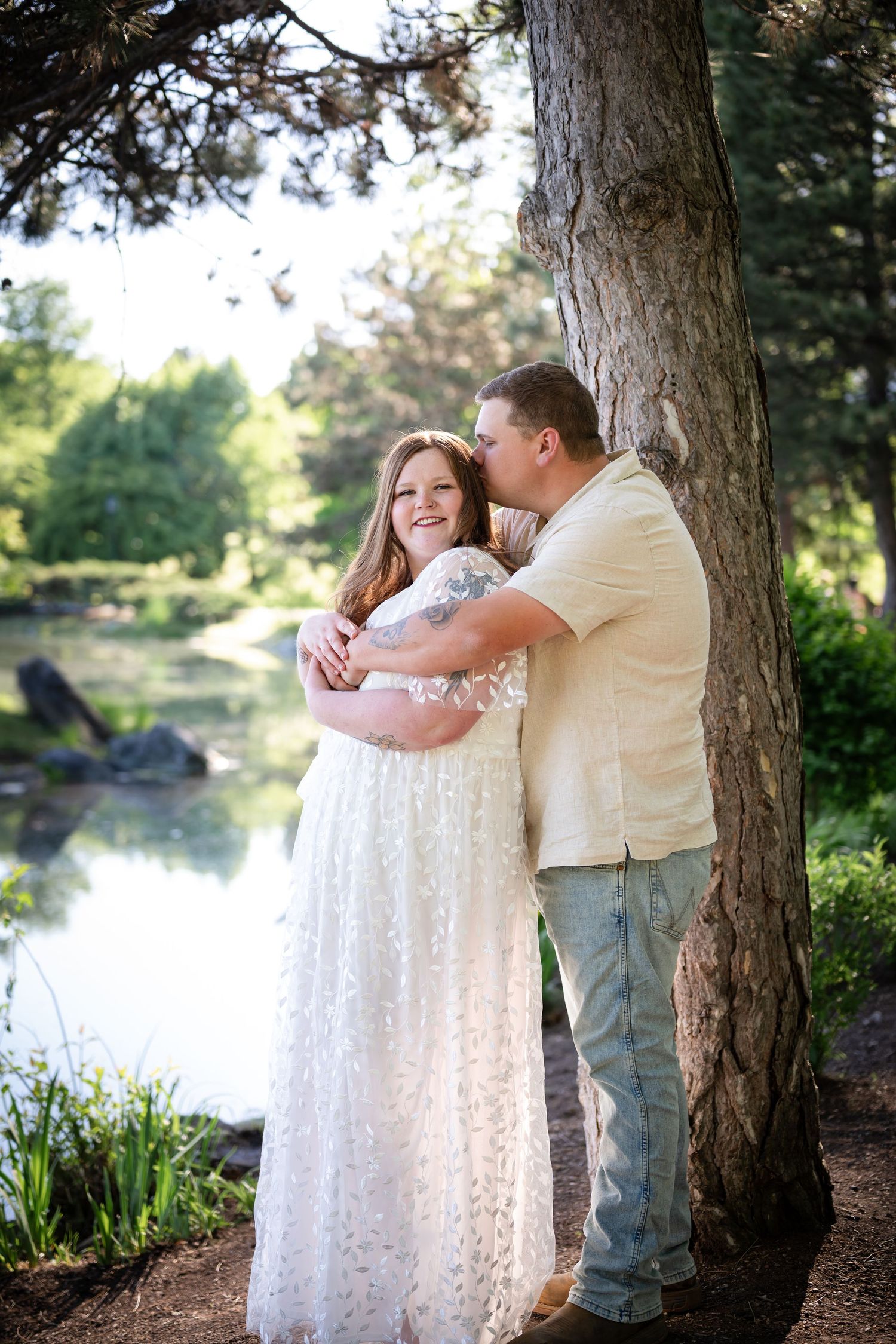 Couple embracing by a tree near a pond, woman in white lace gown, man in beige shirt kissing her cheek outdoors.