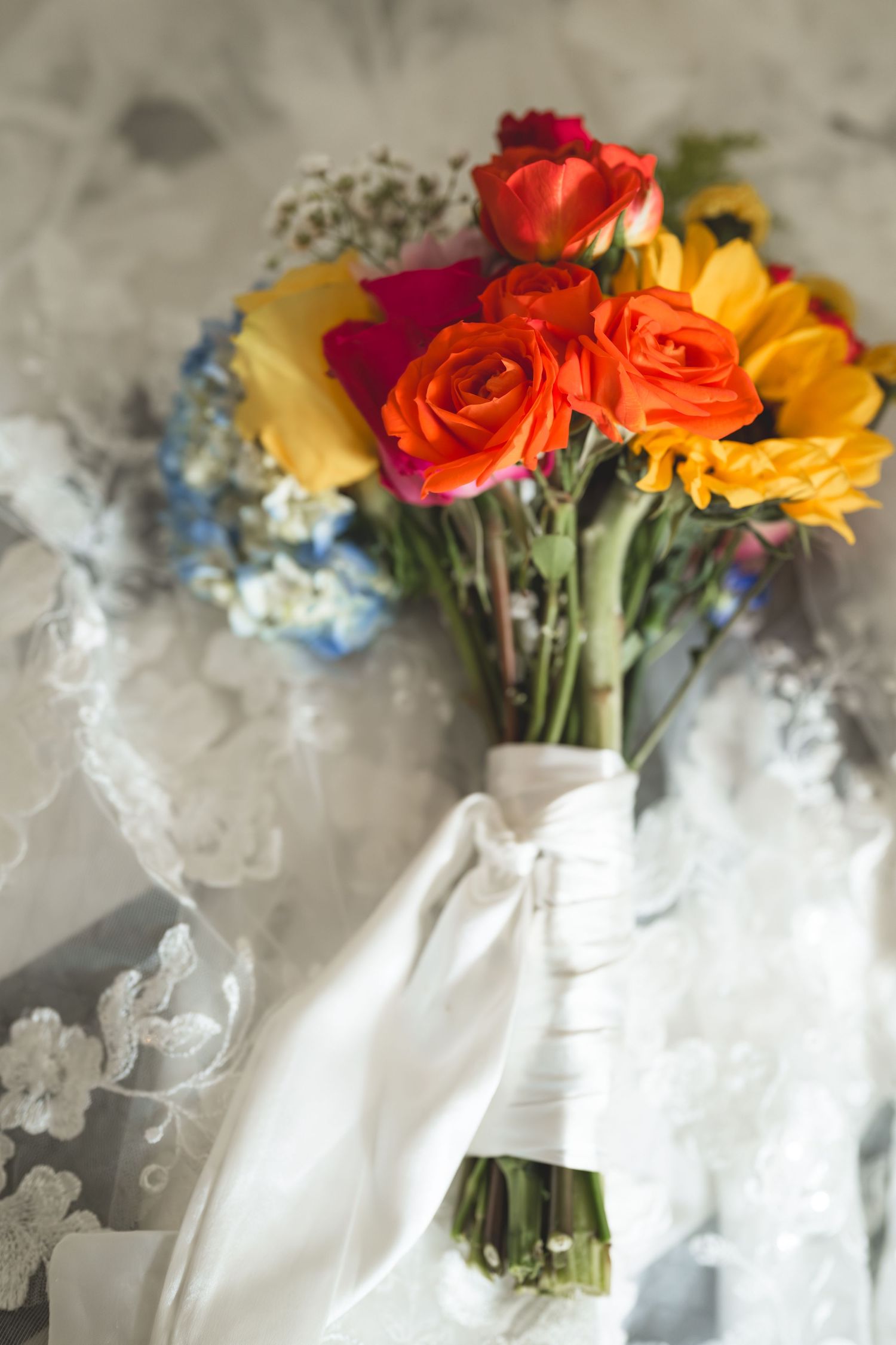 A vibrant bridal bouquet with orange roses, yellow sunflowers, and blue hydrangeas wrapped in white ribbon against lace.