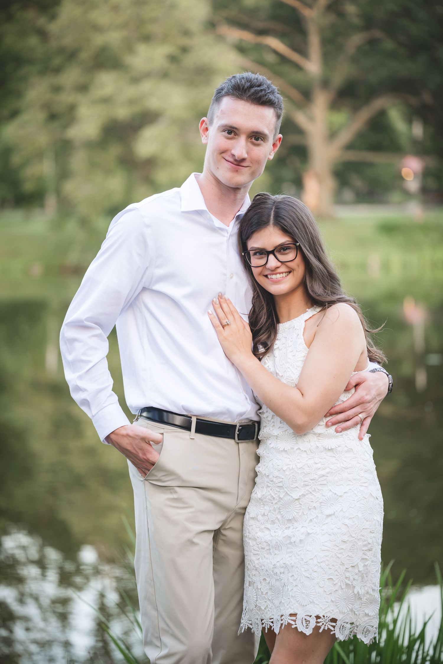 Couple posing outdoors near a pond, man in white shirt and woman in white lace dress with glasses, smiling.
