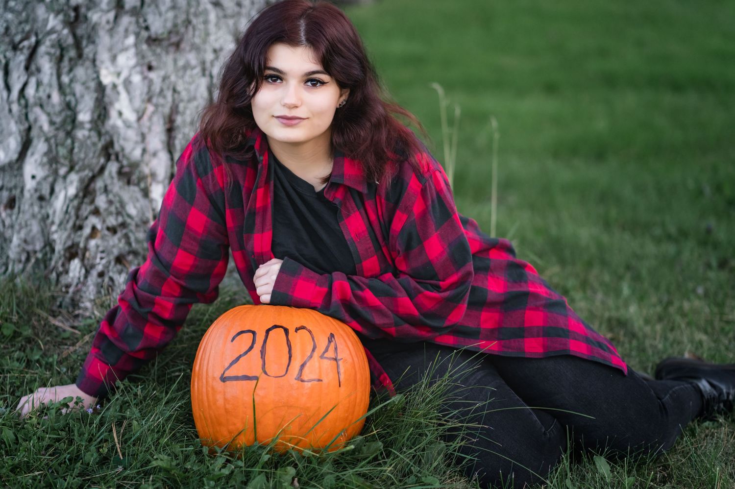 Fall themed portrait with a carved 2024 pumpkin and red plaid flannel by a tree.