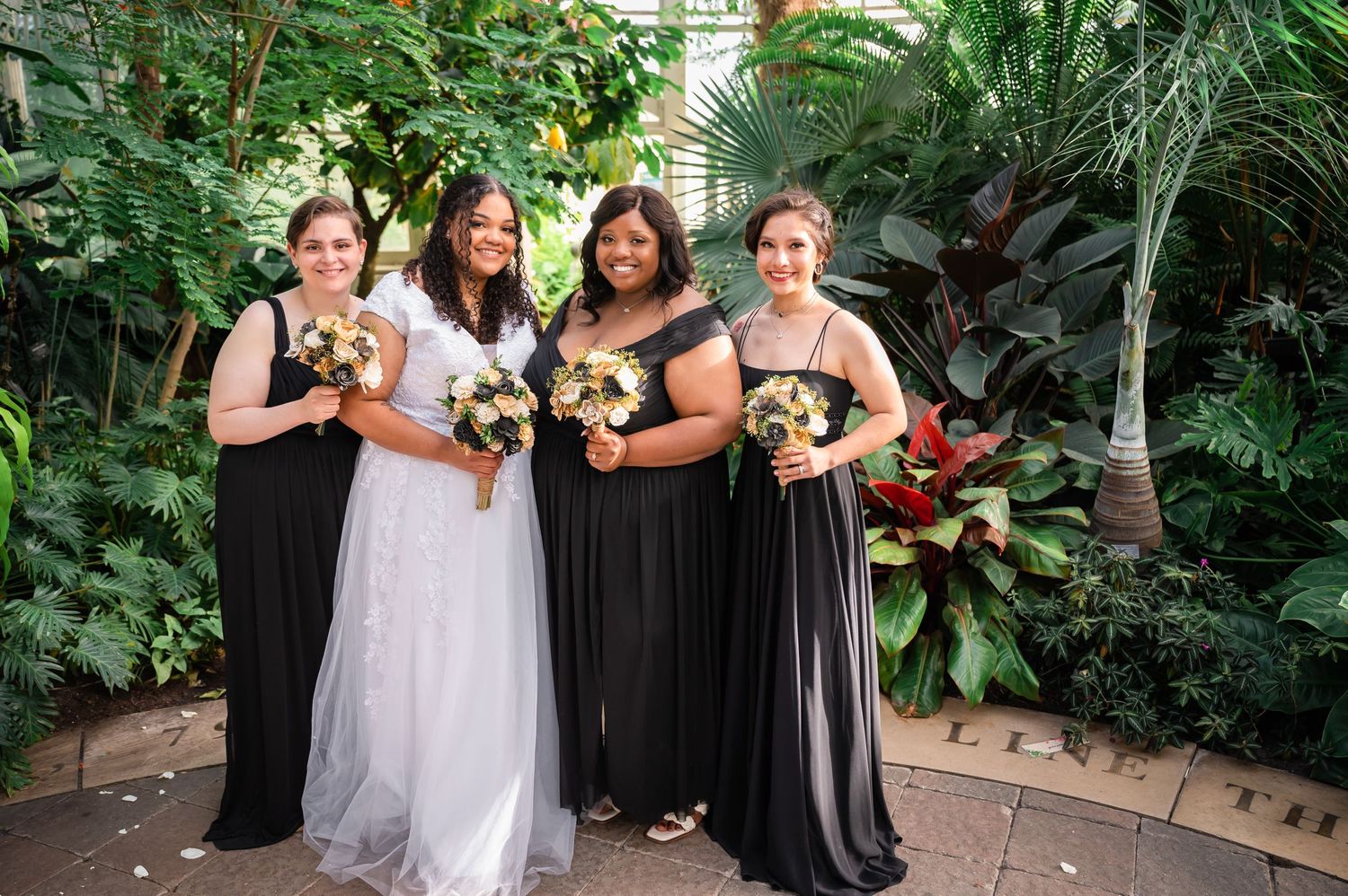 Bridal party with bridesmaids in black dresses pose with bouquets in tropical garden setting.