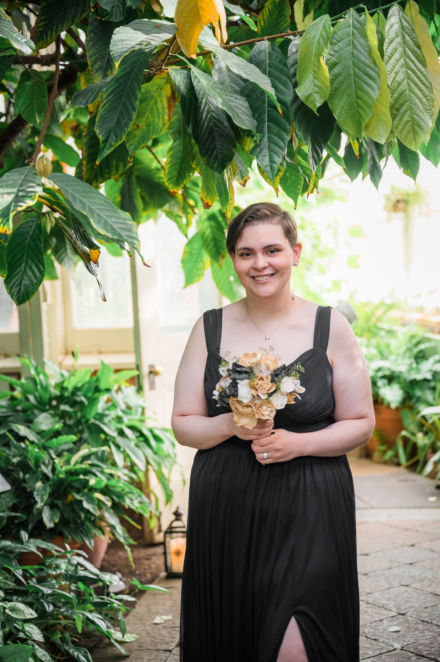 Person in black formal dress holds a peach and cream floral bouquet under tropical foliage.