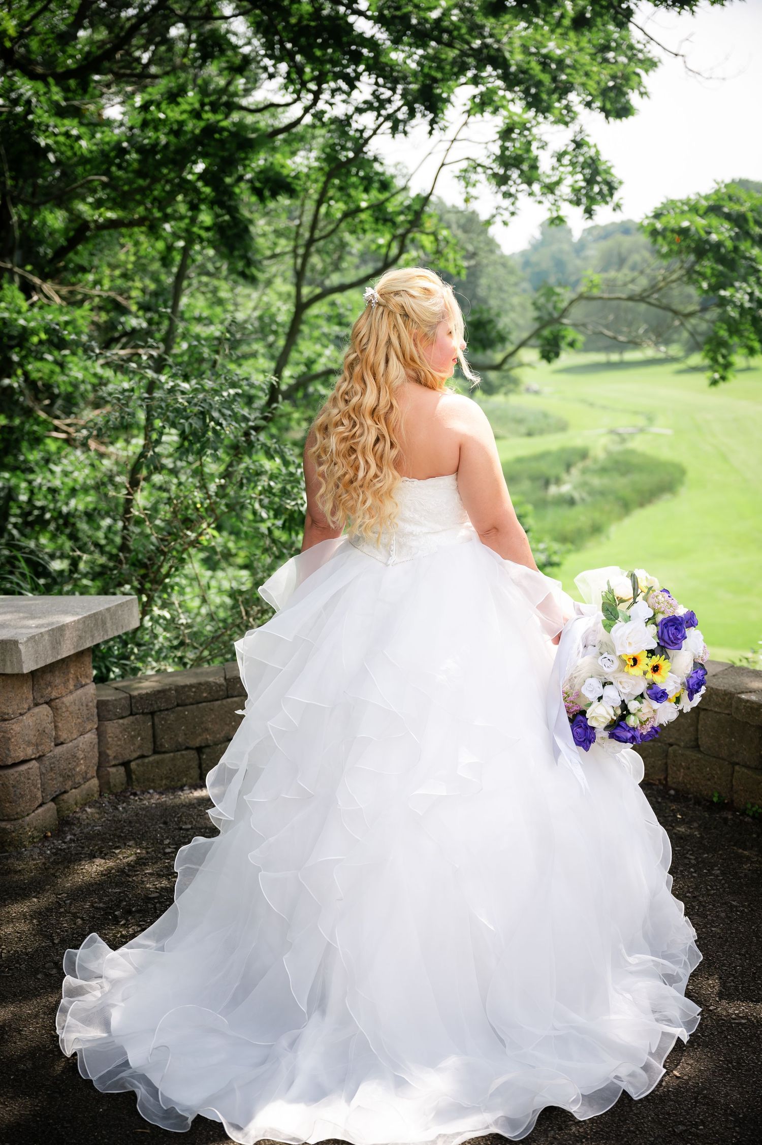 A bride in a layered tulle ballgown holds a purple and white bouquet while overlooking a scenic landscape.