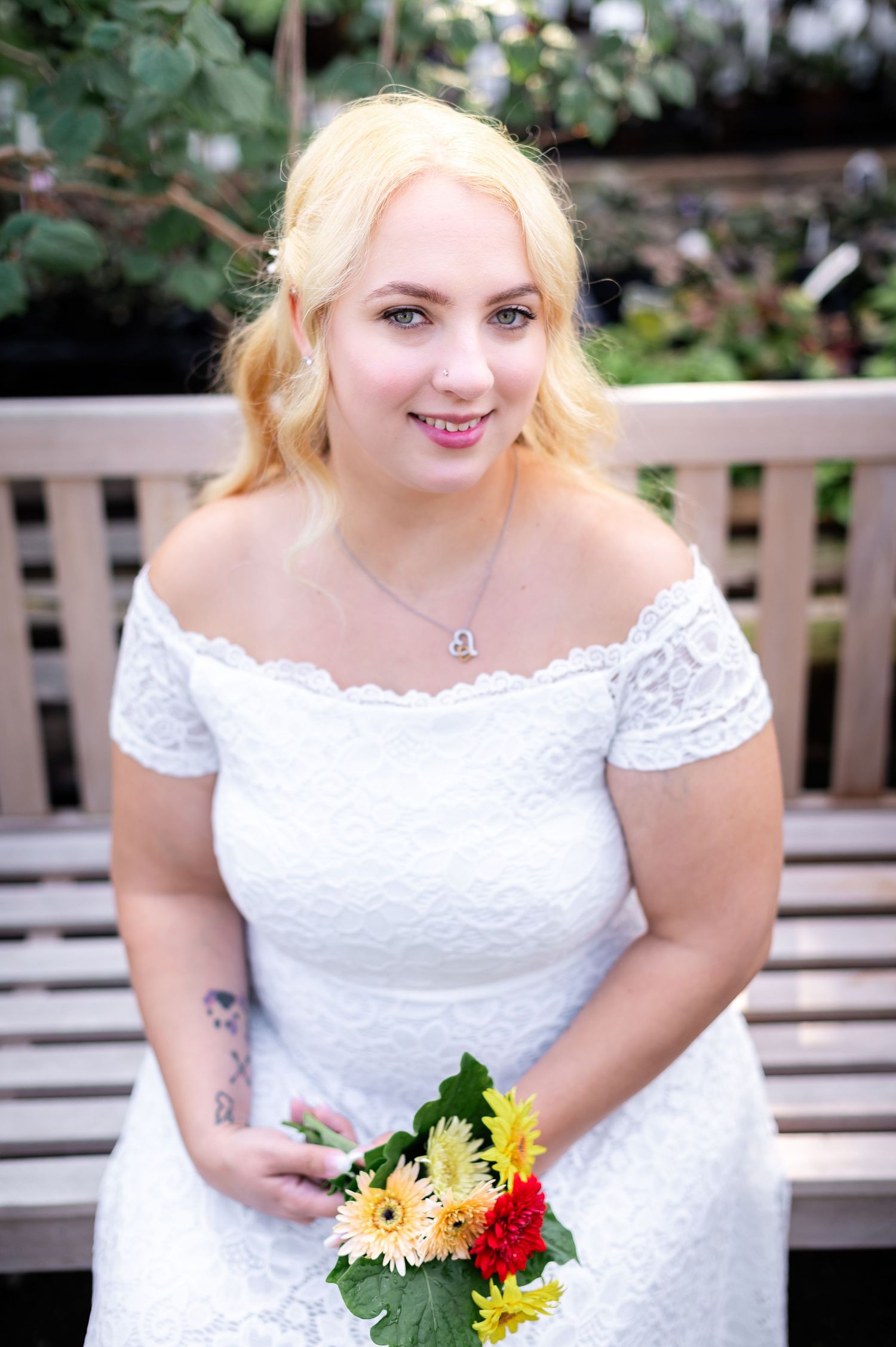 A bride in a white lace off-shoulder dress holds a colorful bouquet while sitting on a garden bench.