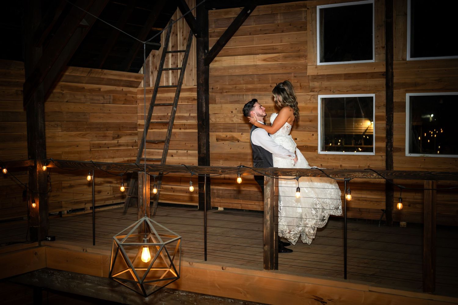 A romantic nighttime scene inside a rustic barn with warm lighting and wooden walls, featuring a couple in wedding attire.