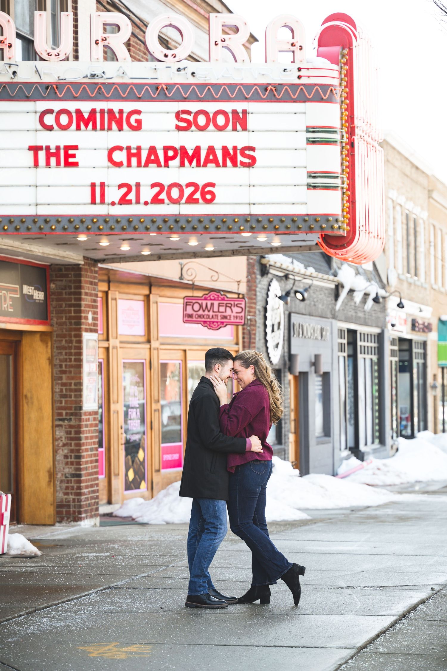Engaged couple embracing outside Aurora Theatre with wedding announcement marquee sign.