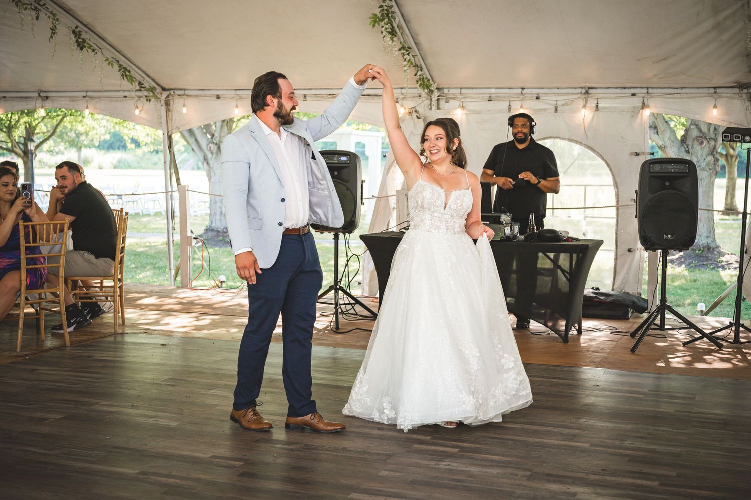 Bride and groom share first dance under white tent at outdoor wedding reception with DJ in background.