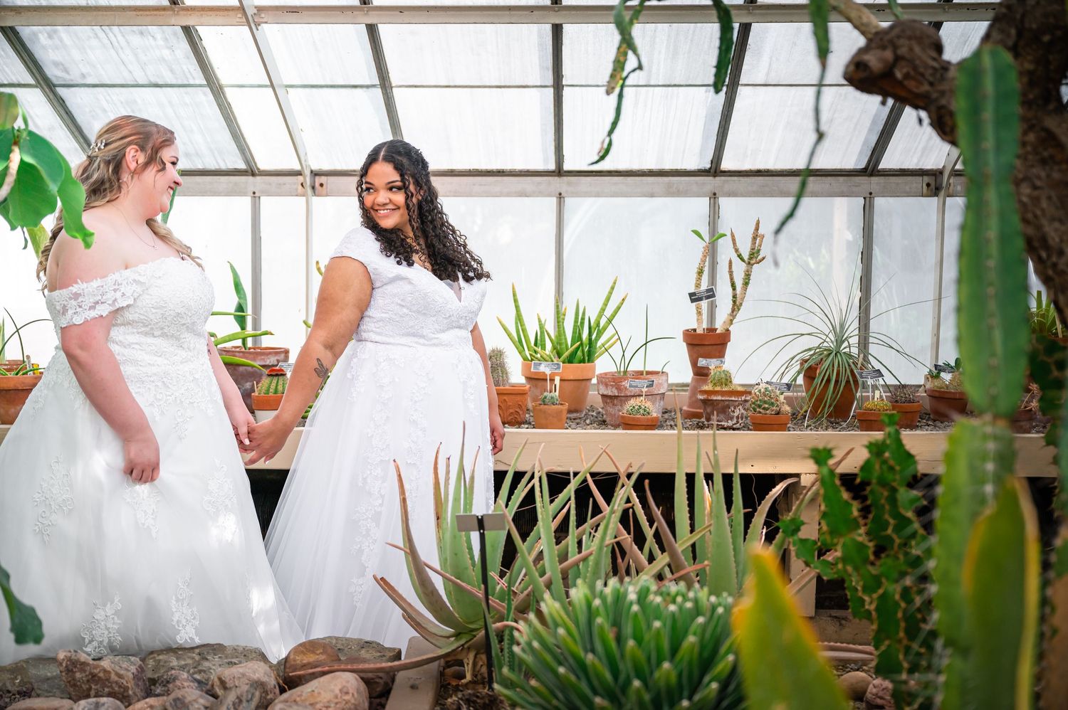 Two brides in white gowns stand together in a greenhouse filled with tropical plants and natural light.