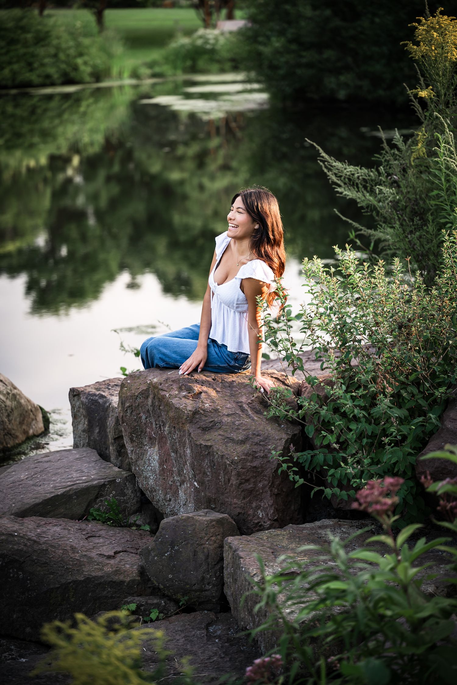 Woman in white dress sitting on rocks by serene pond surrounded by greenery.