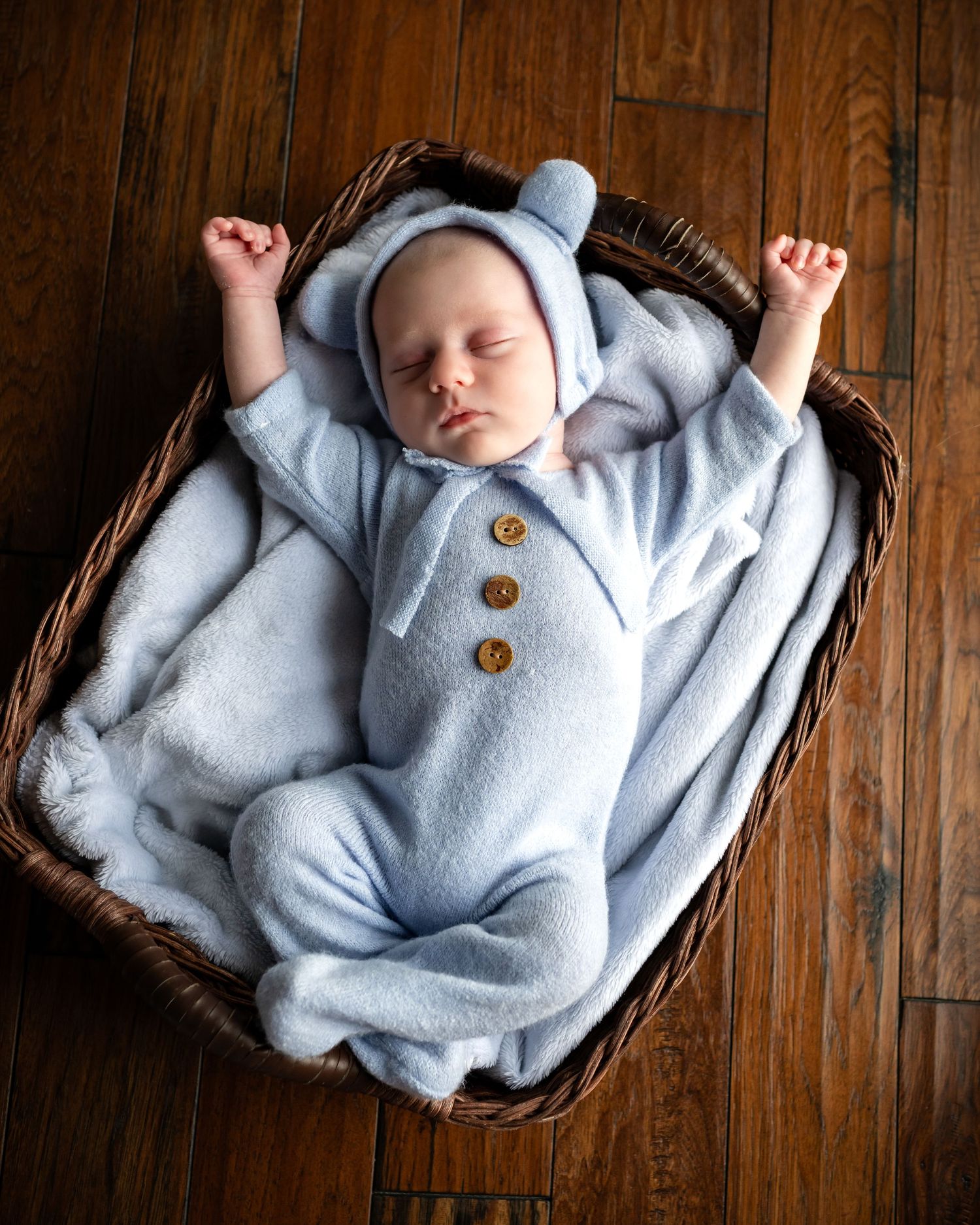 Sleeping baby wearing soft blue onesie with buttons lies peacefully in wicker basket on hardwood floor.