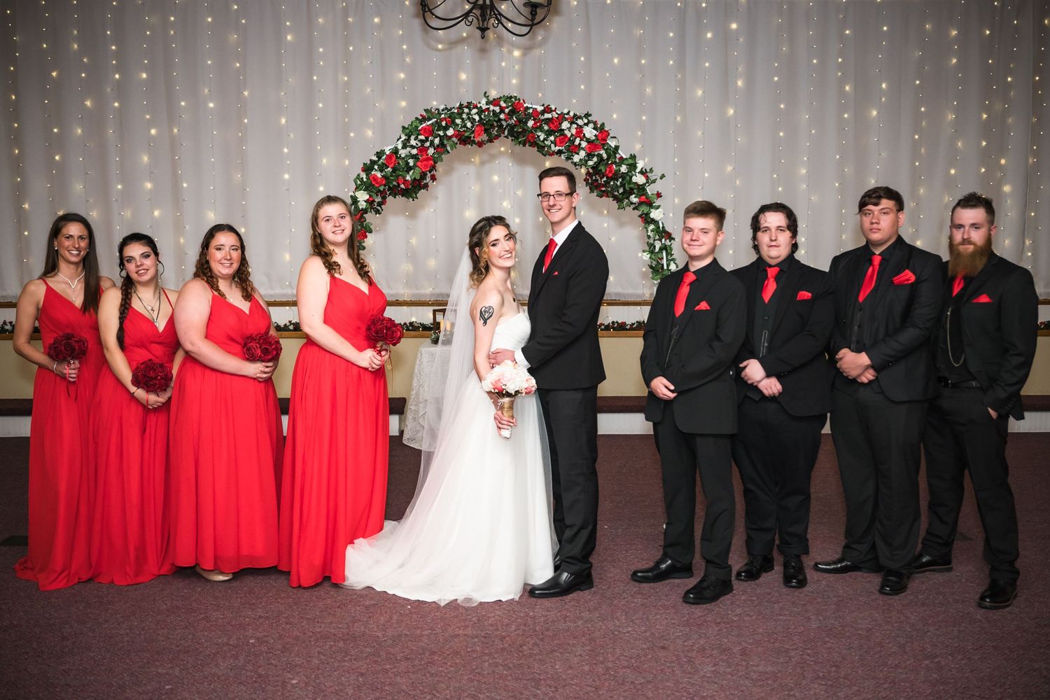Wedding party lined up with bridesmaids in red dresses and groomsmen in black suits under a decorated arch.