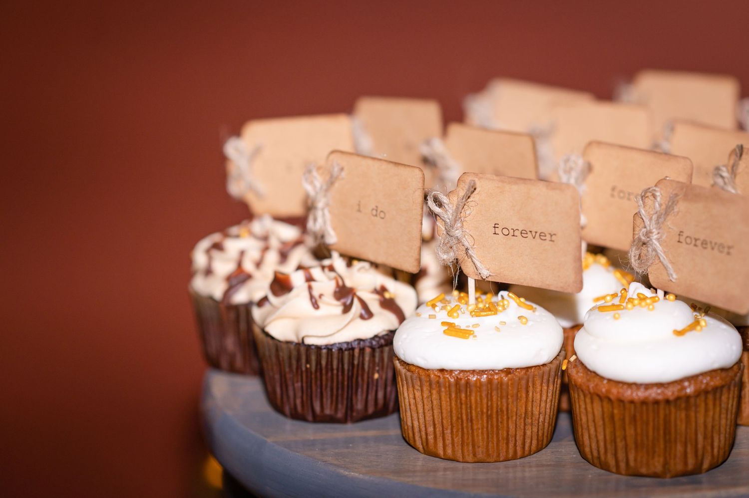 Elegant wedding cupcakes with name cards displayed on a blue stand against a burgundy background.