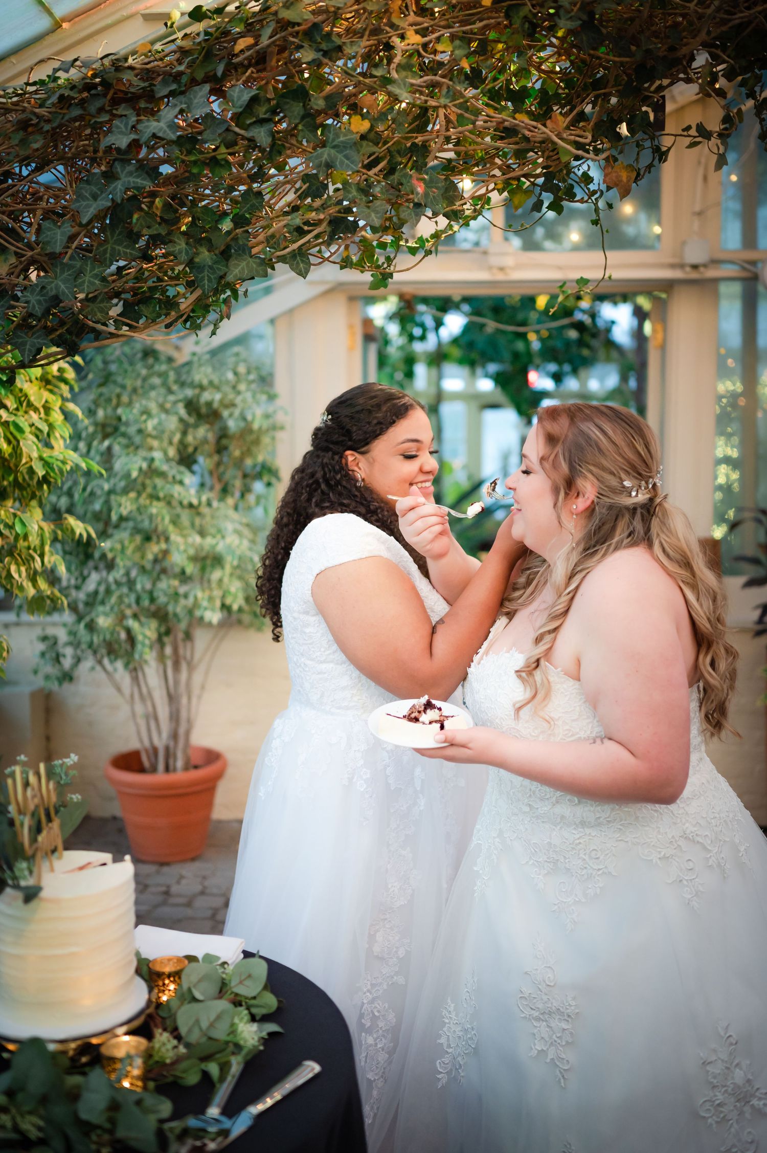 Two brides share wedding cake during intimate garden reception moment.