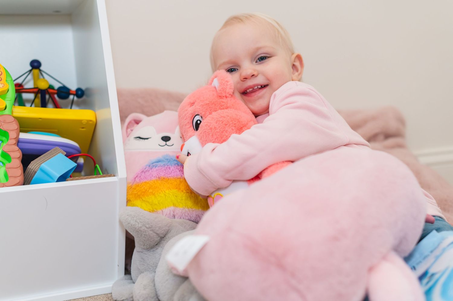 Baby in pink clothing hugging a colorful plush toy while sitting by toy shelf.