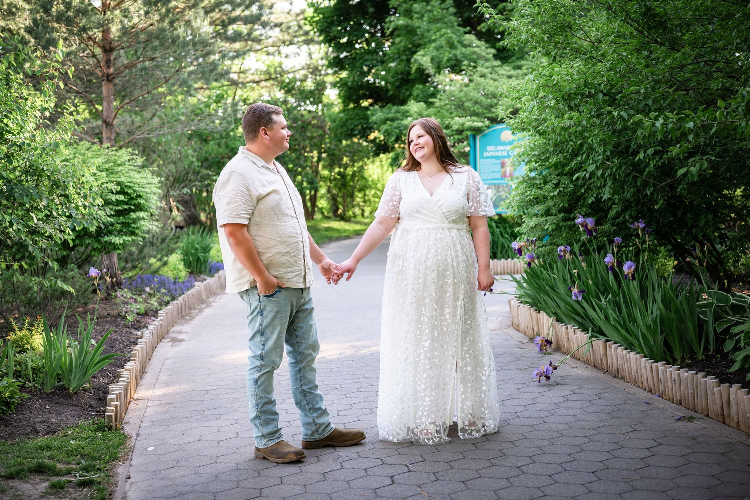 Couple holding hands on a brick garden path surrounded by lush greenery, woman in white lace gown, man in casual attire.