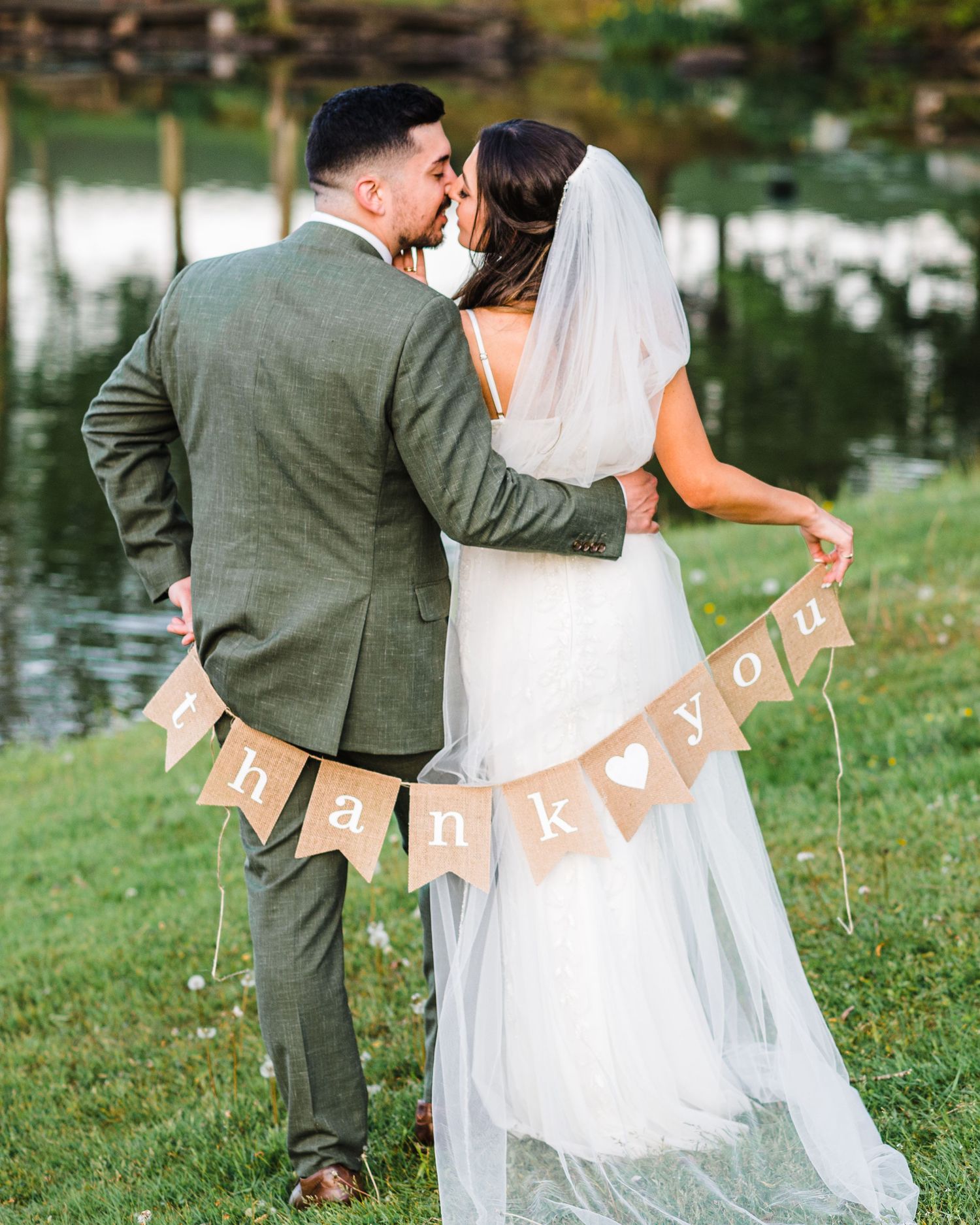 A couple standing by a lake sharing a romantic kiss with a kraft paper 'I LOVE YOU' banner displayed behind them.