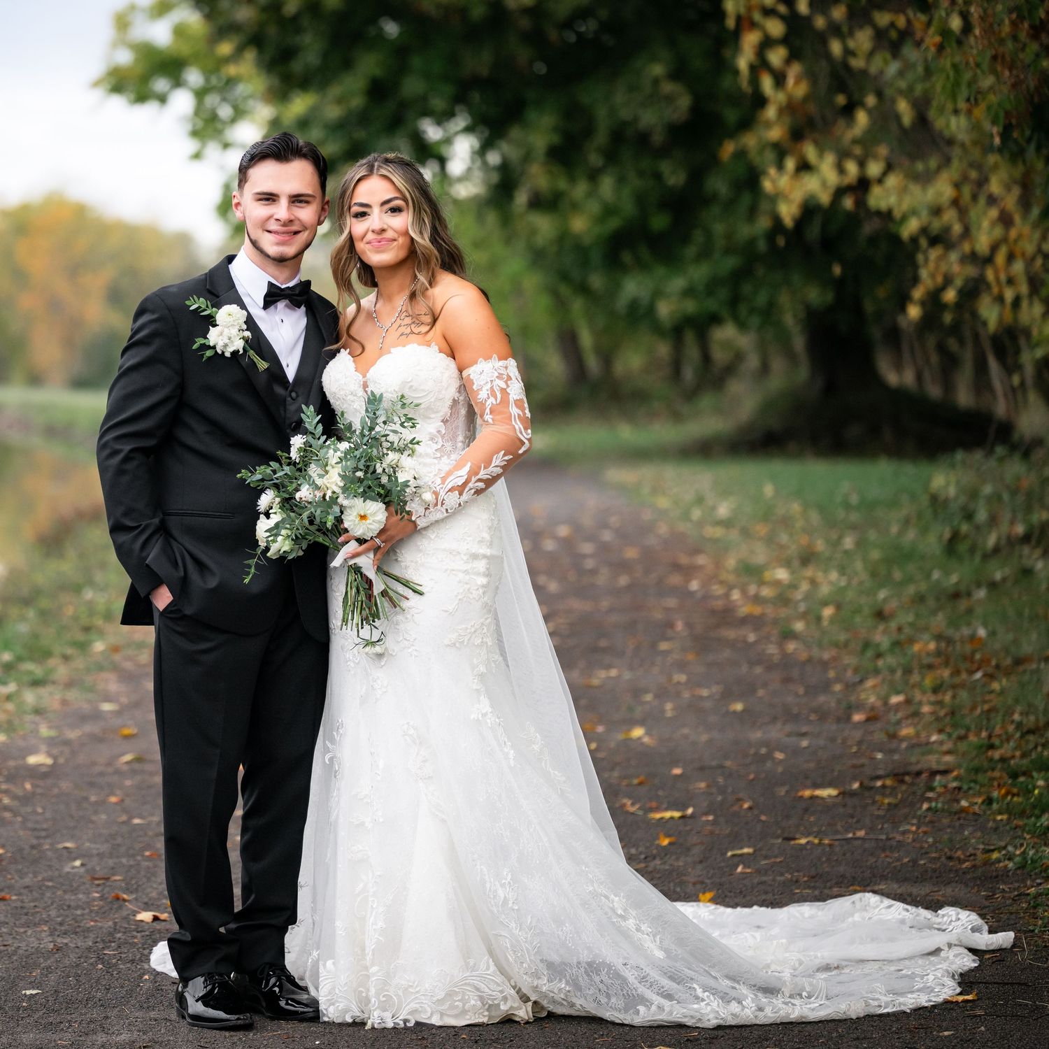 A couple in wedding attire pose together on a tree-lined path covered in fallen autumn leaves during their special day.