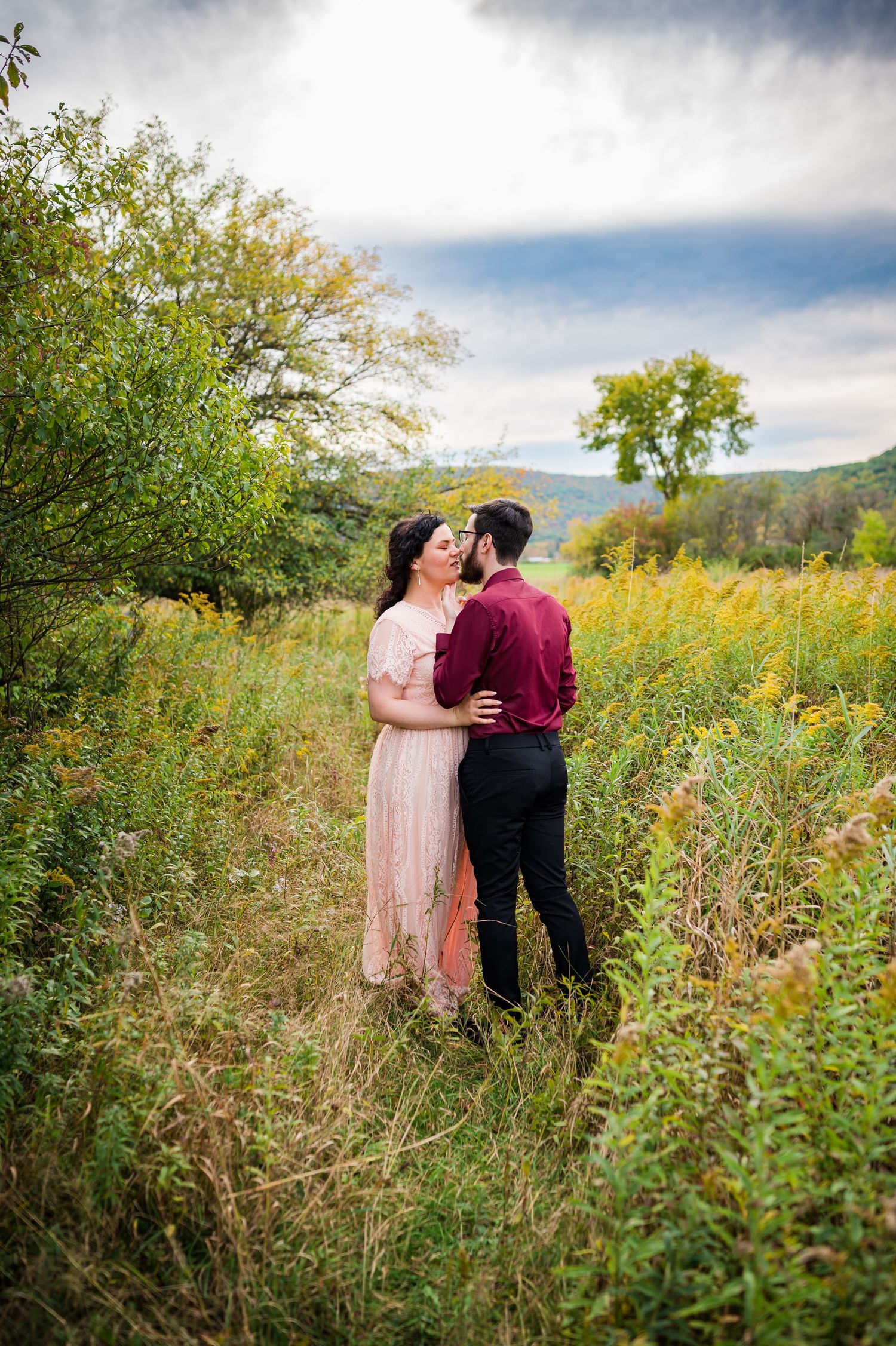 A romantic couple embraces in a wildflower meadow with mountains and dramatic clouds in the background.