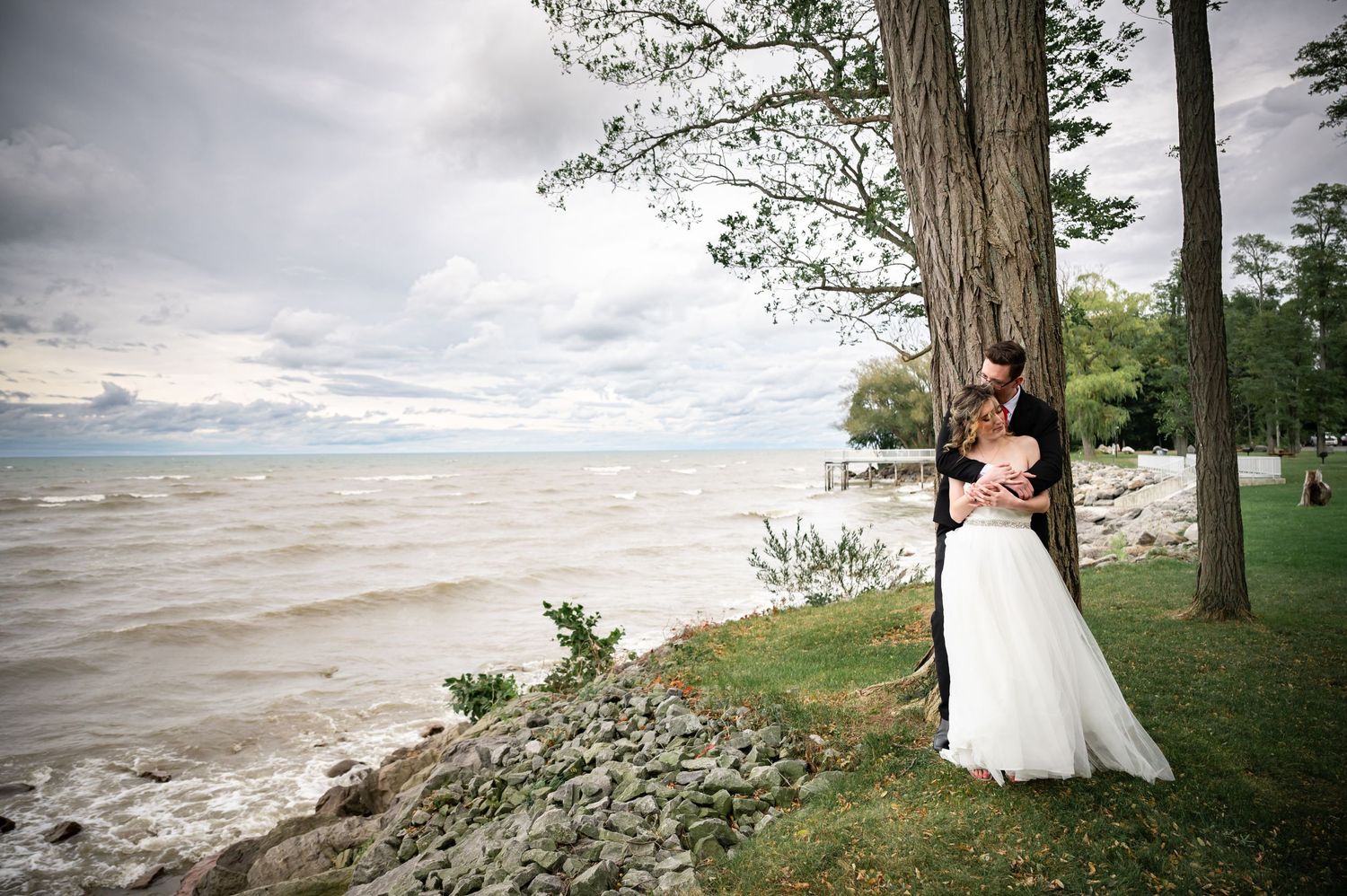 Romantic waterfront wedding photo with couple embracing on rocky shoreline under cloudy skies.