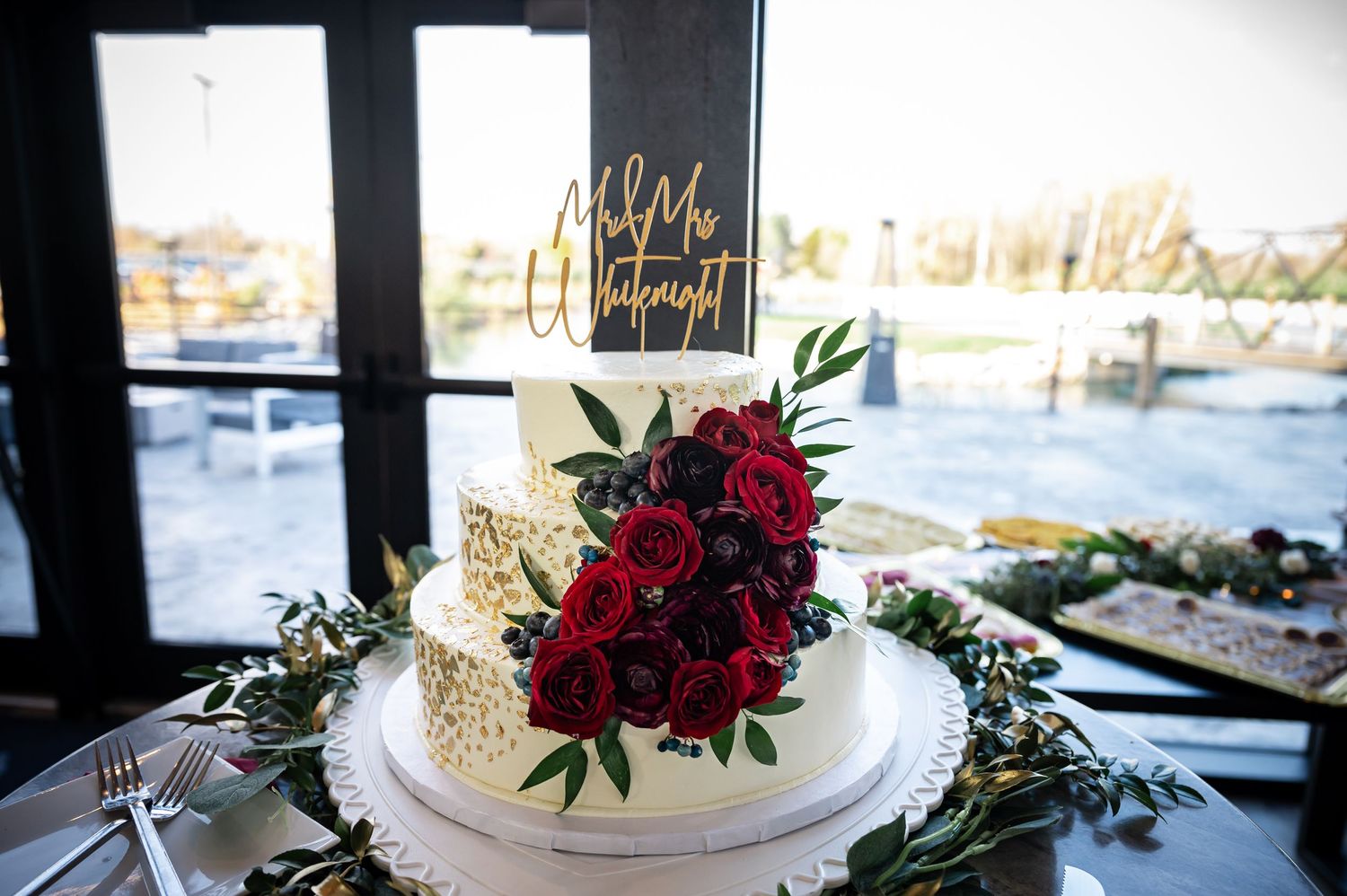 White wedding cake decorated with deep red roses and greenery against window backdrop.