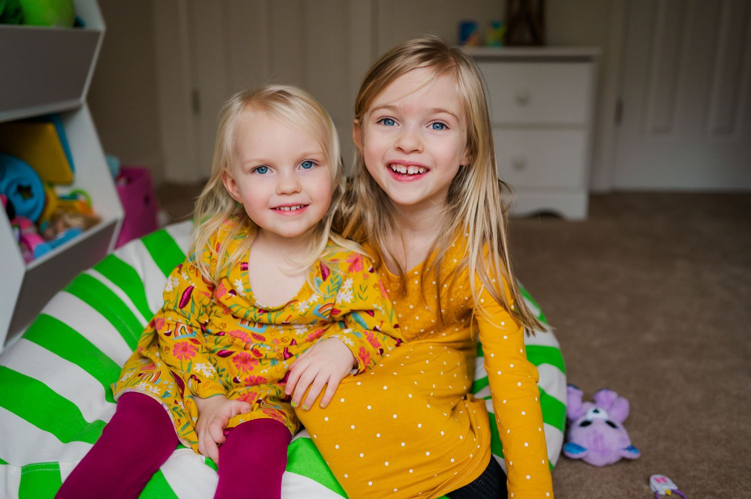 Two children in bright yellow floral dresses sitting together on a green and white striped chair.