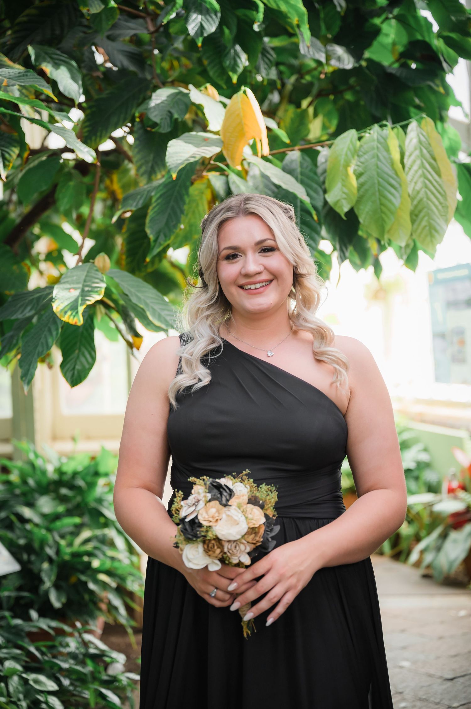Elegant black one-shoulder dress styled in a garden conservatory with tropical foliage.