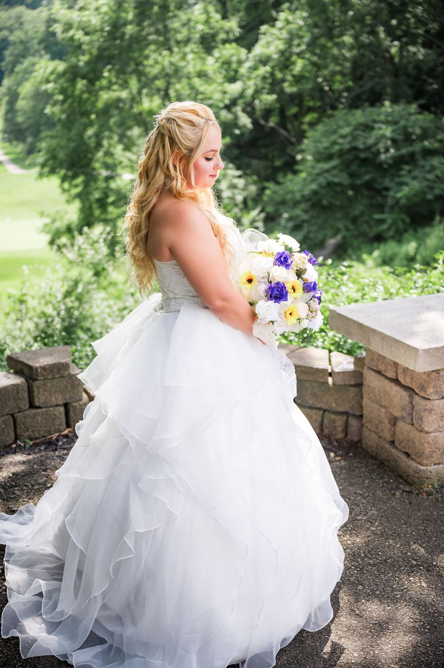 Side view of bride holding purple and yellow bouquet while standing on stone overlook.