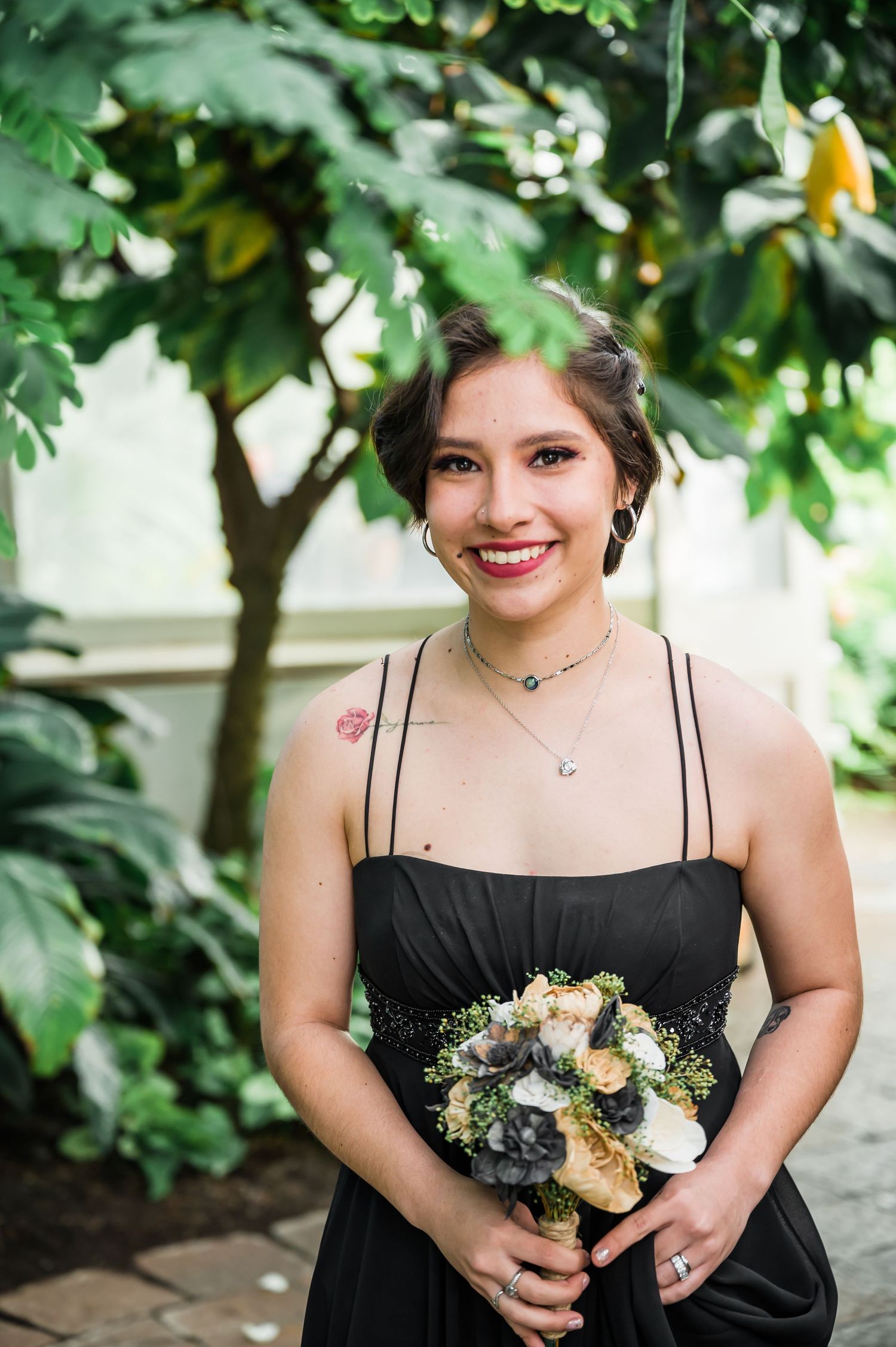 Person in black formal dress holds floral bouquet while standing among green foliage.