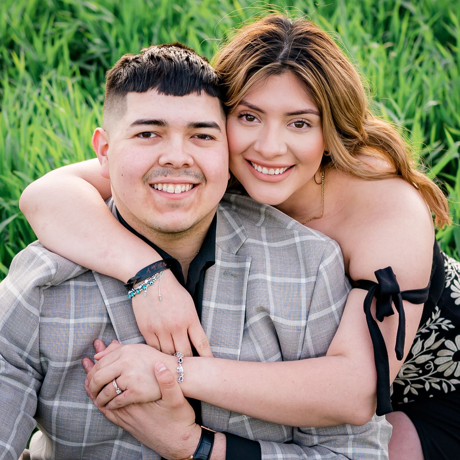 A couple in coordinated grey outfits embrace and smile during an outdoor photoshoot in tall green grass.