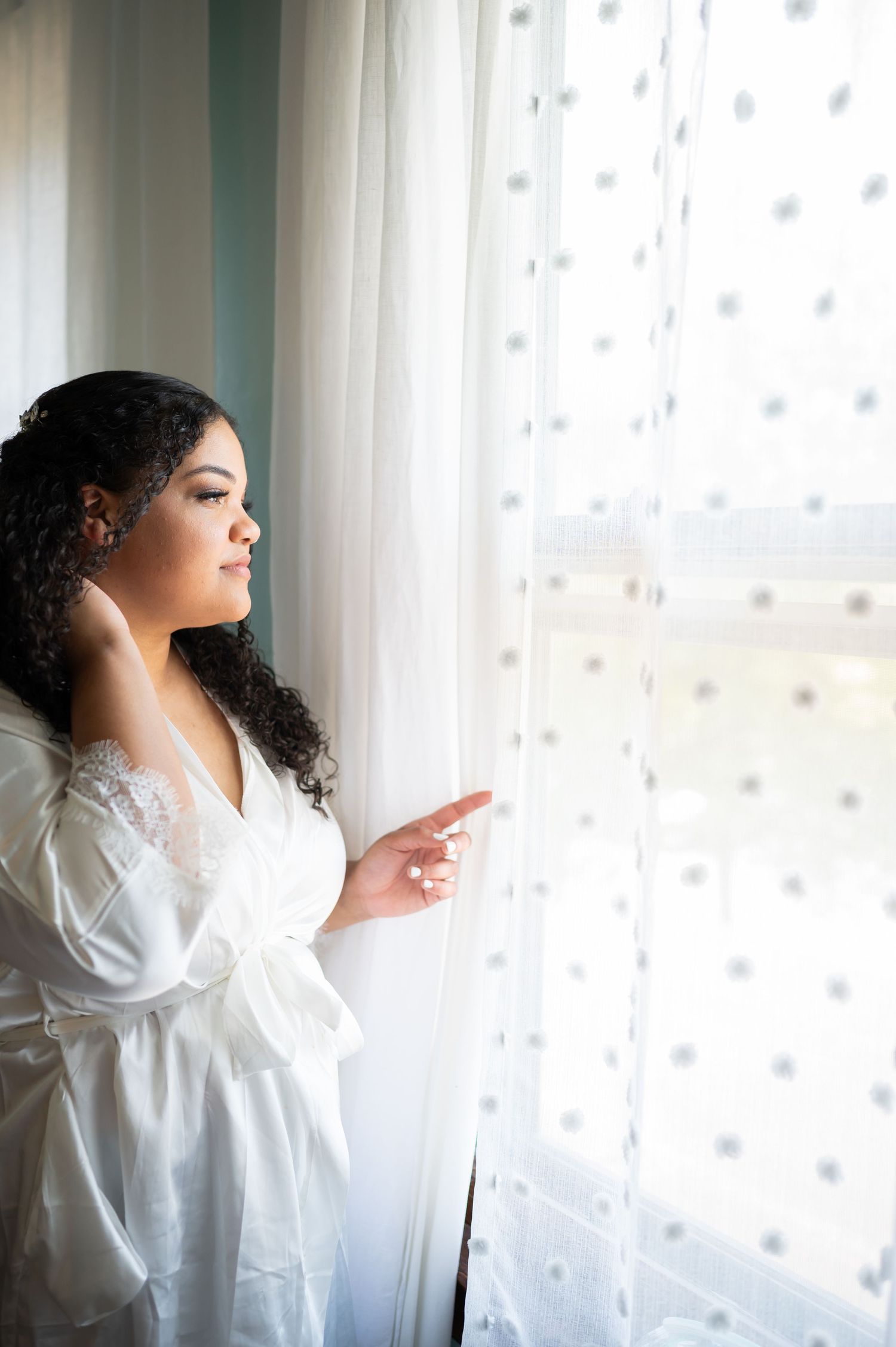 A woman in a white robe gazes thoughtfully through a sheer curtained window.