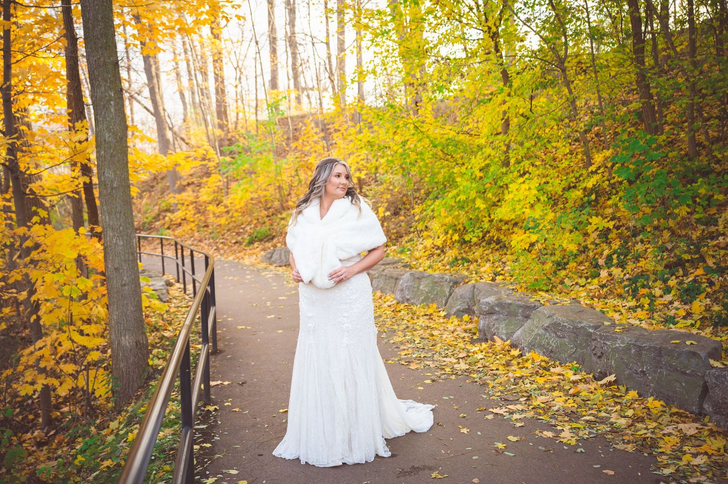Bride in flowing white gown walks along autumn path surrounded by golden leaves.